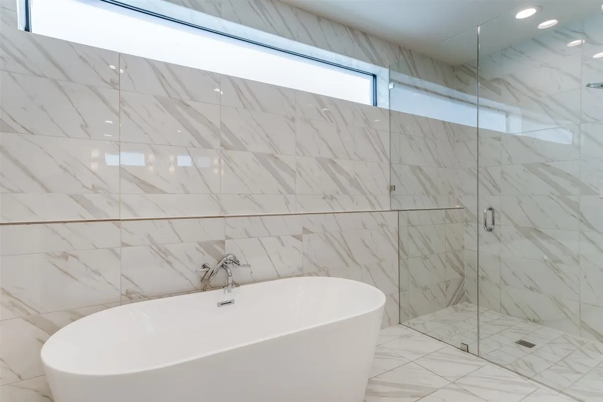Freestanding soaking tub with chrome fixtures, glass-enclosed shower, and full-height tiled walls featuring subtle veining