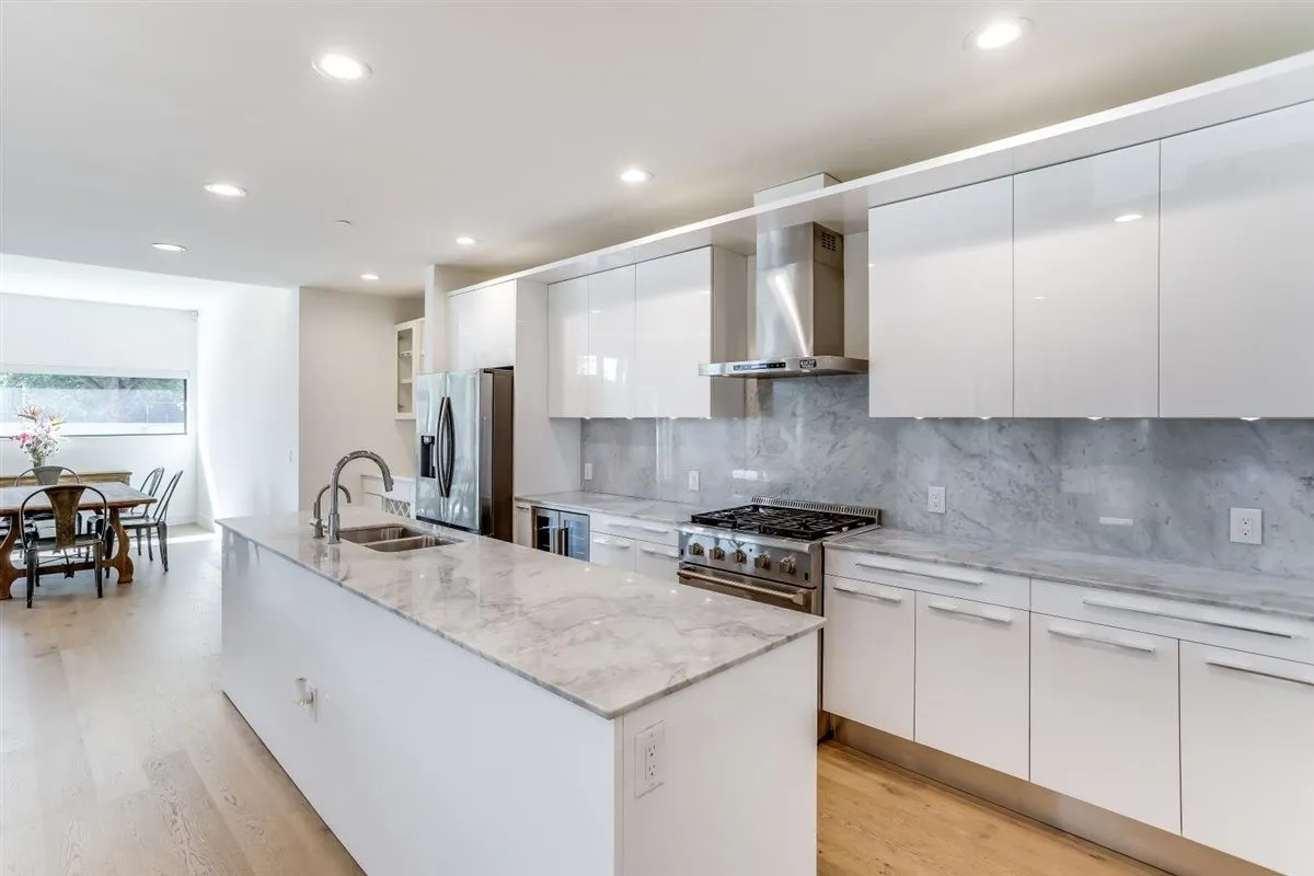 Modern kitchen featuring a large island with sink, stainless steel appliances, white flat-panel cabinetry, and light wood-finish flooring
