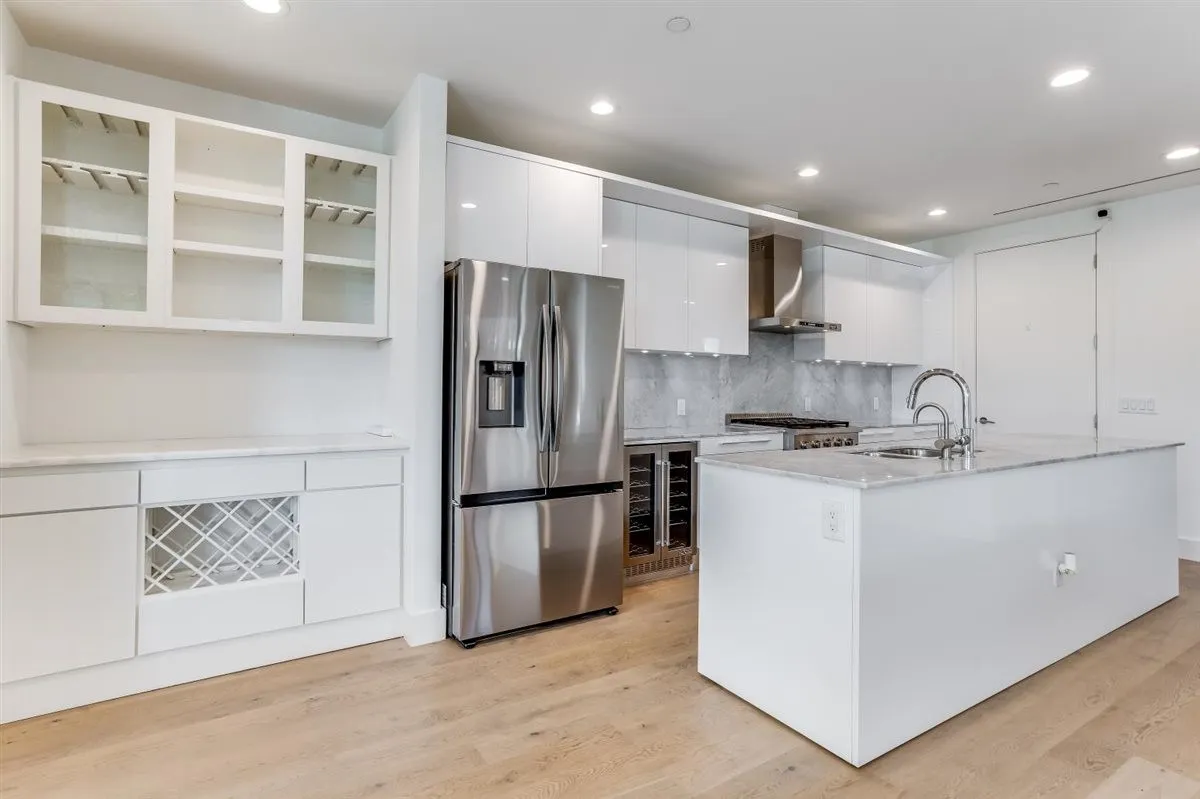 Modern kitchen featuring a large center island with integrated sink, stainless steel appliances, abundant white cabinetry, light wood-finish flooring, and recessed lighting