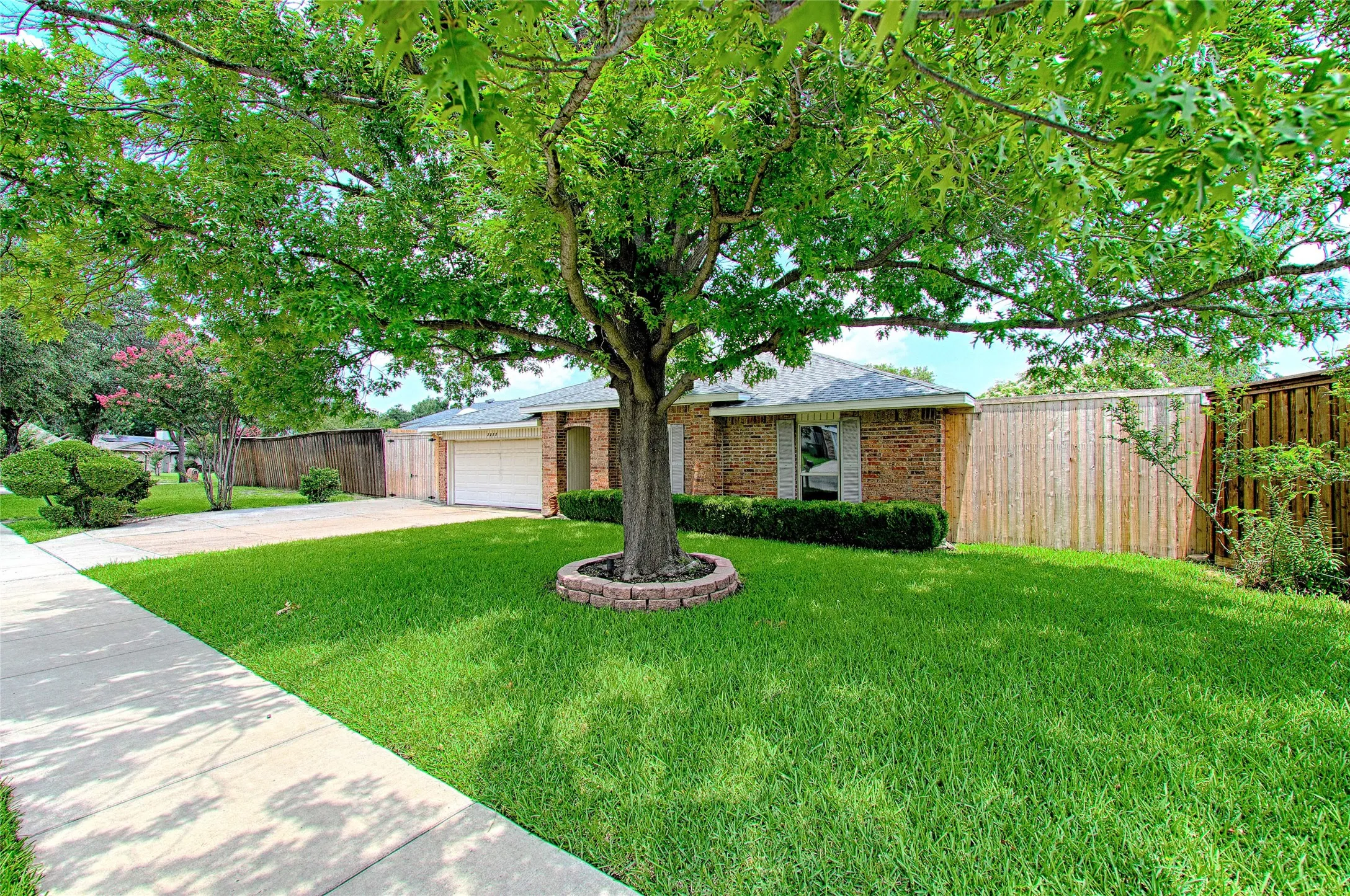 Front exterior featuring a brick facade, attached two-car garage, mature shade tree, manicured lawn, and privacy fencing