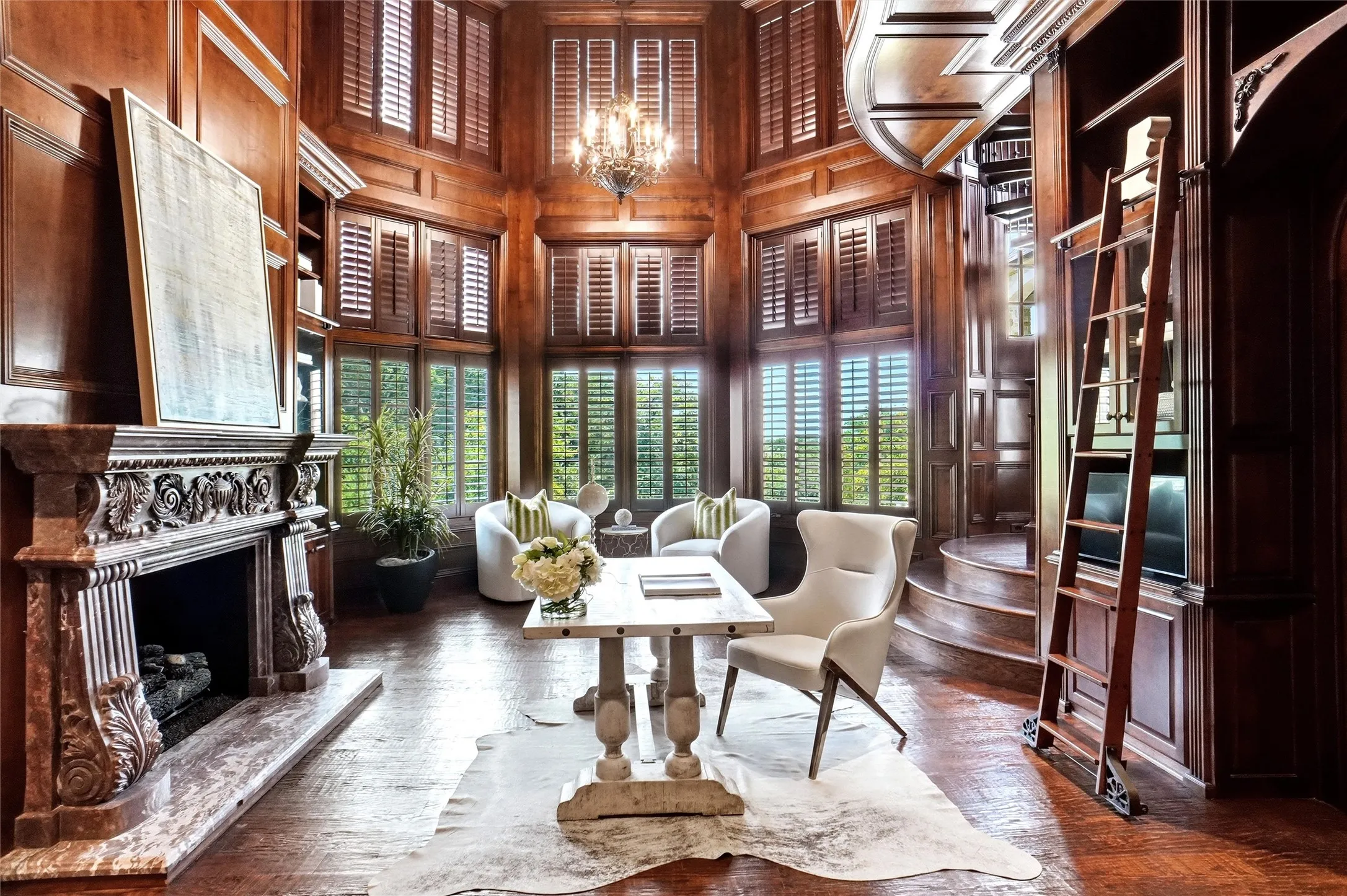Hardwood flooring throughout featuring a carved stone fireplace with gas logs, built-in shelving, panel walls, and multiple windows with plantation shutters. There is a spiral staircase off the sittling area that leads to the second level.