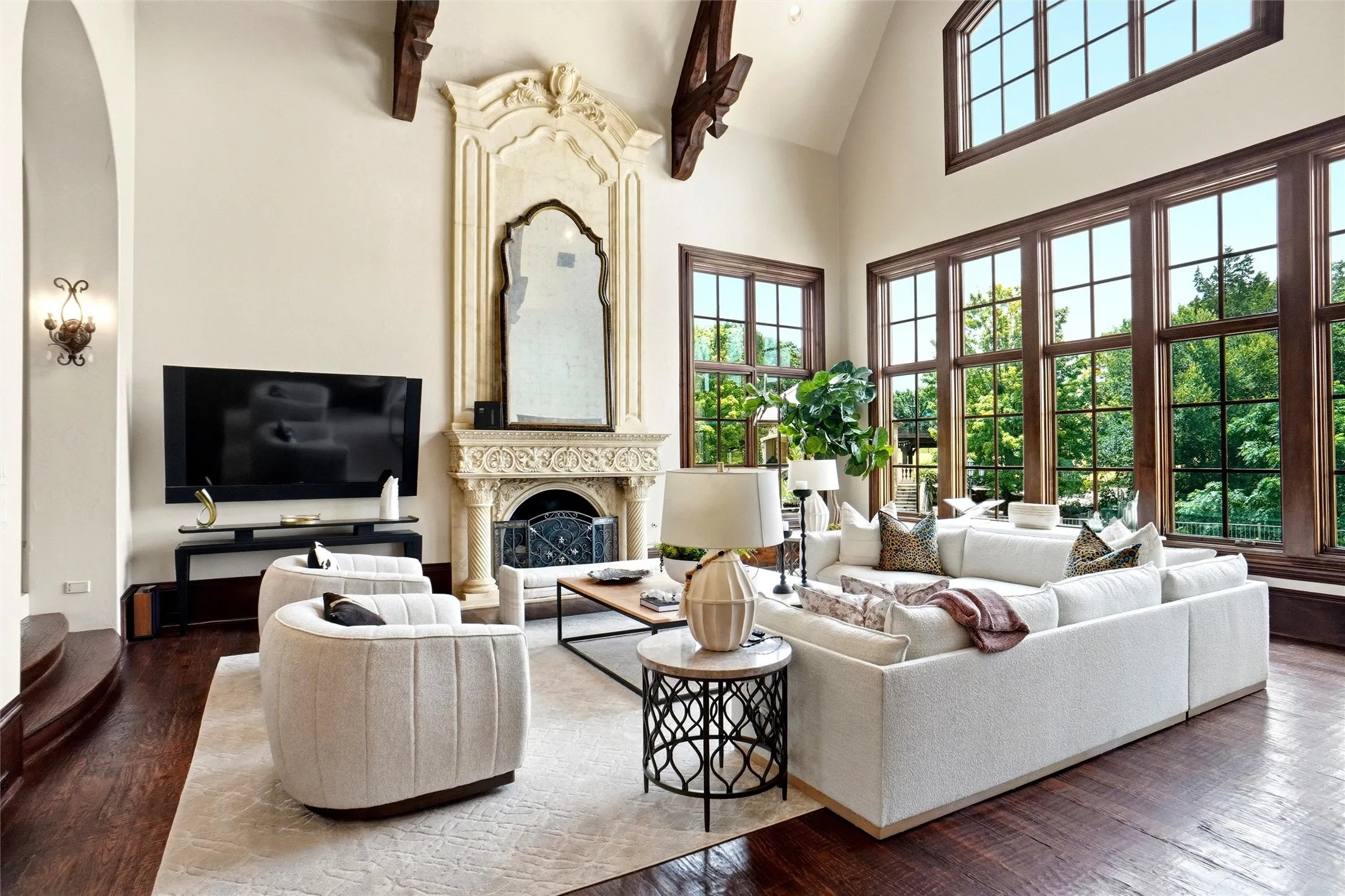Vaulted ceiling living area featuring a carved stone fireplace with an ornate mirror, extensive dark-frame windows, exposed architectural ceiling beams, and rich wood-finish flooring