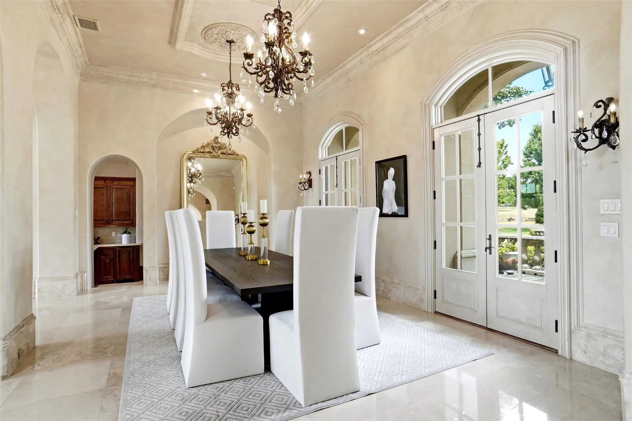 Formal dining area featuring coffered ceilings with two ornate chandeliers, light-toned stone flooring, and arched French doors with transom windows