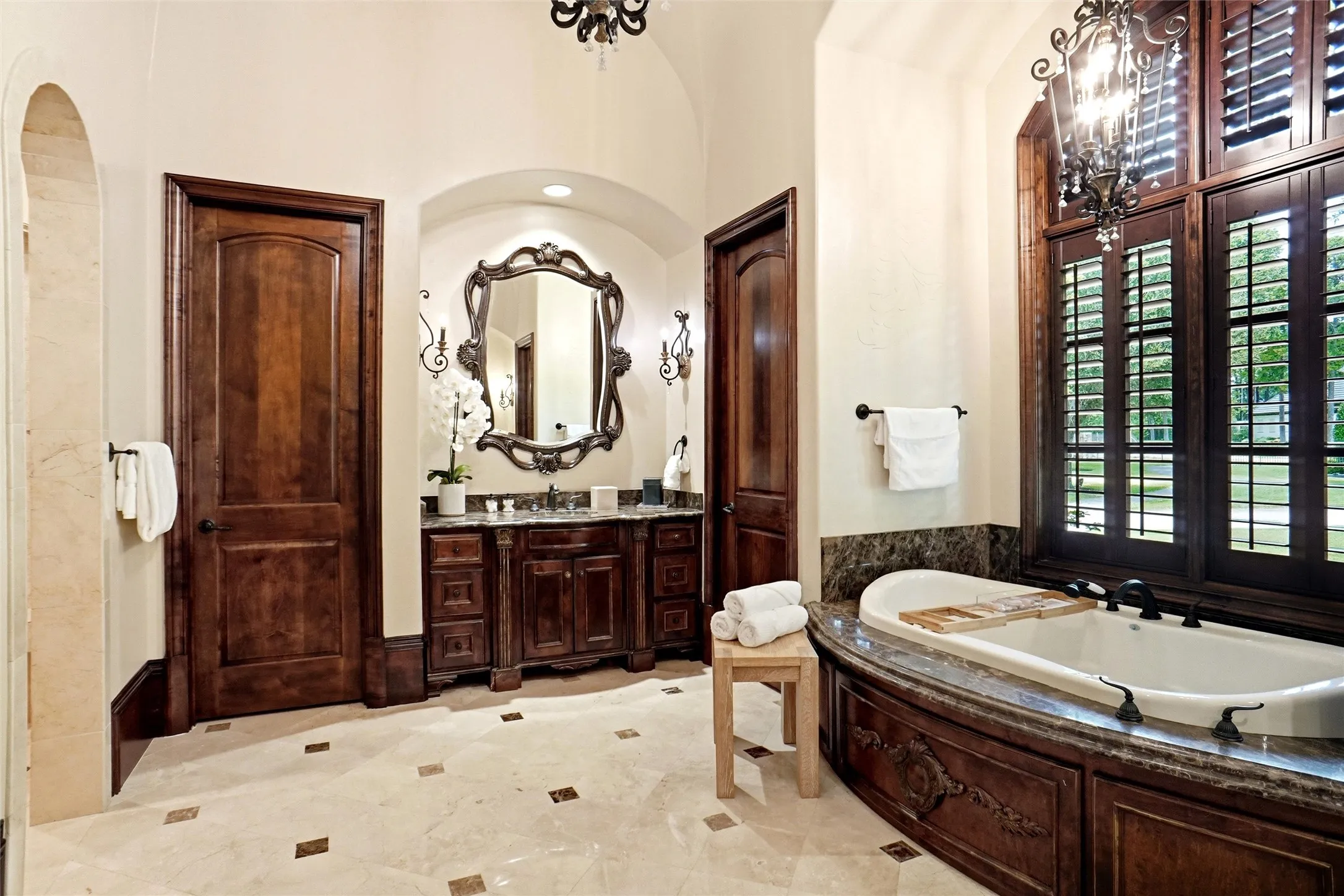 Spacious bath featuring an oval soaking tub with dark stone surround, intricate wood paneling, and a large multi-pane window with interior plantation shutters