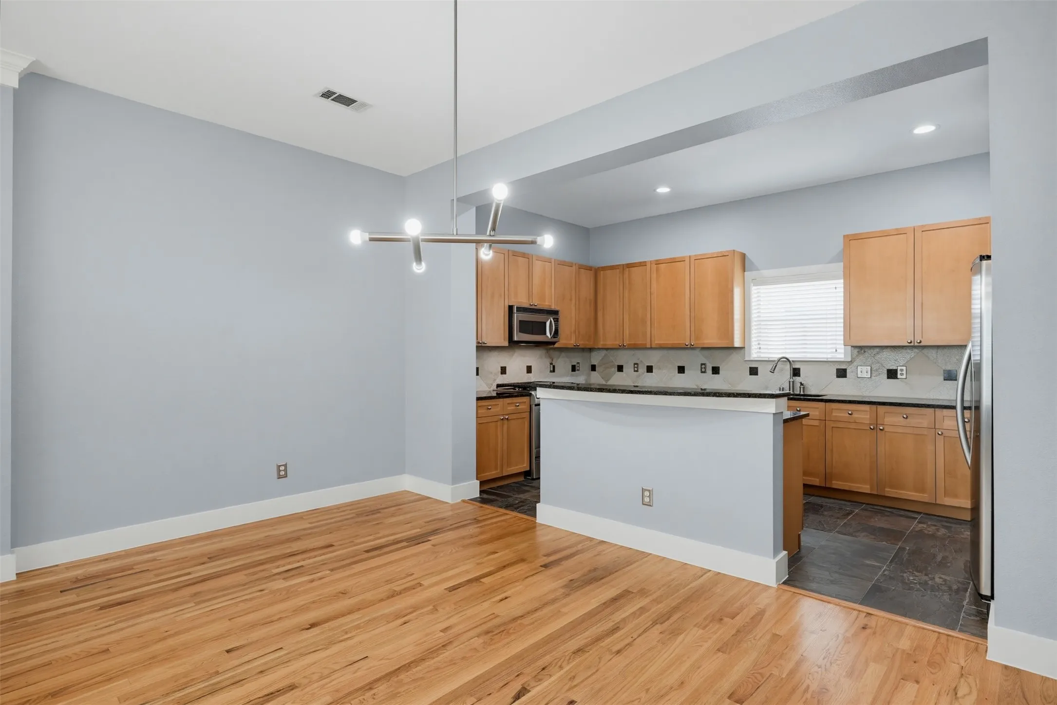 Open-concept living area featuring wood-finish flooring, a contemporary linear chandelier, and a kitchen with wood cabinetry, stainless steel appliances, and a tiled backsplash