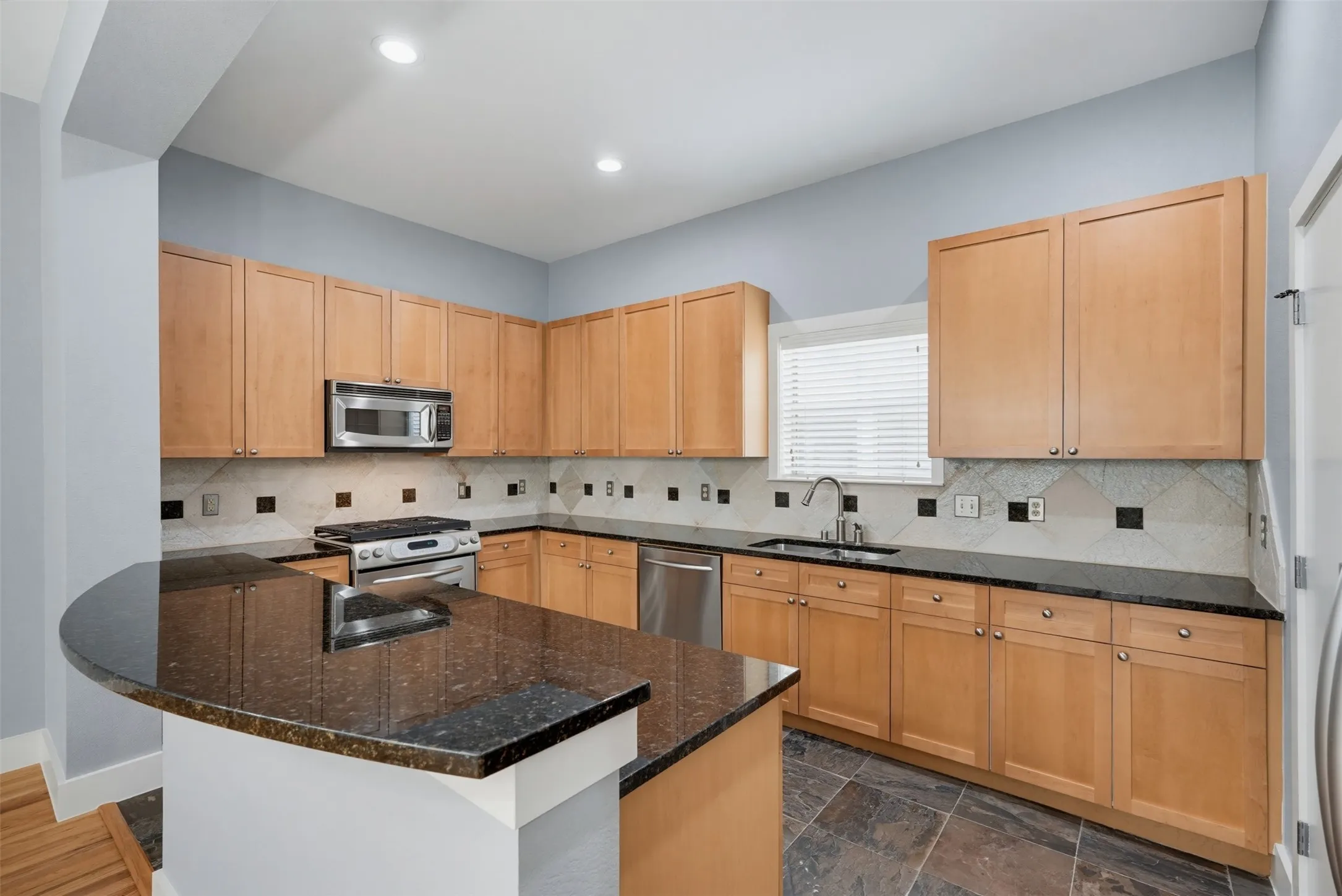Kitchen featuring a curved peninsula with a dark countertop, light wood cabinetry, a stainless steel microwave, a stainless steel range, and a stainless steel dishwasher