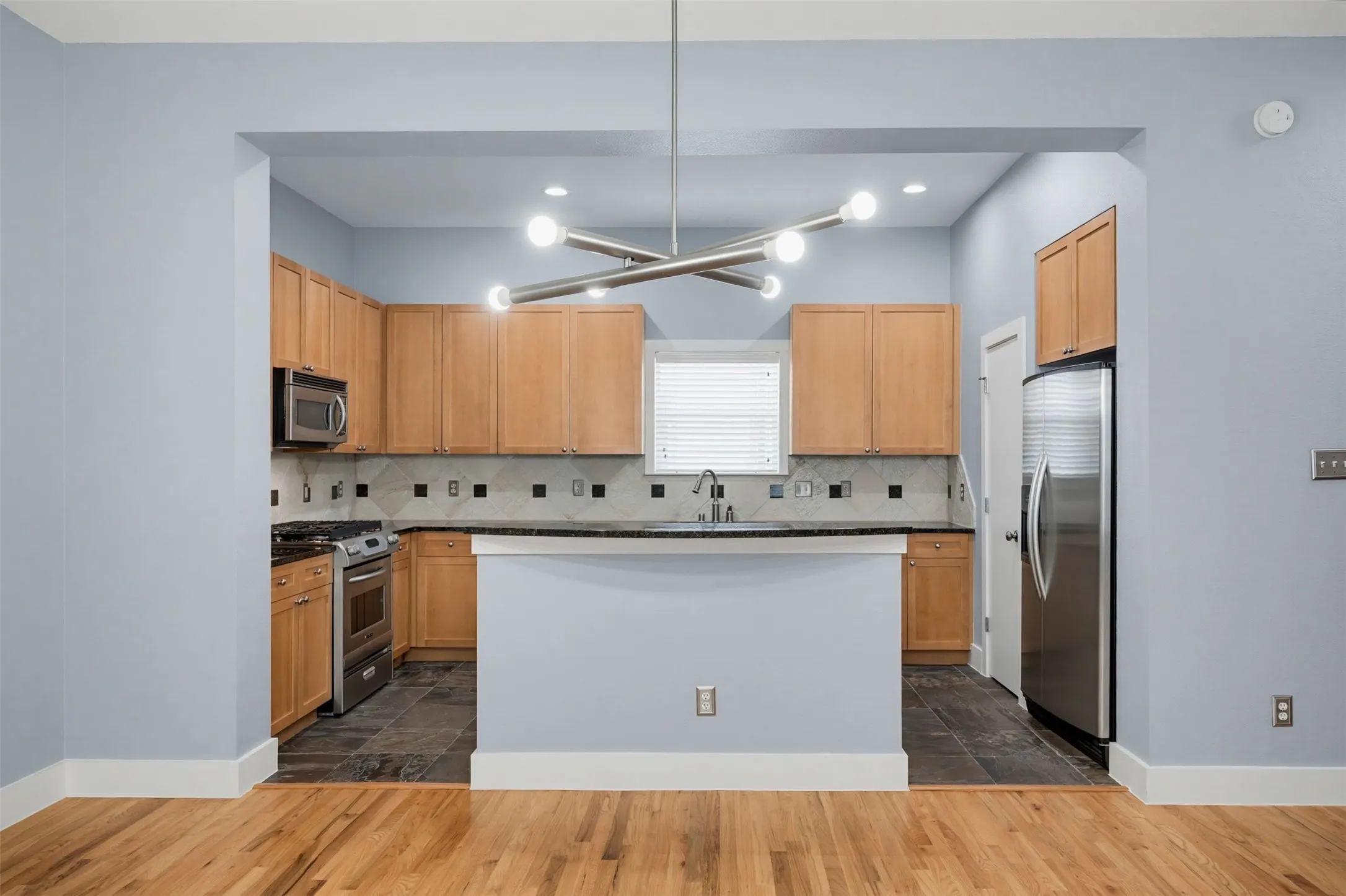 Kitchen with natural wood cabinetry, stainless steel appliances, dark countertops, and a tiled backsplash