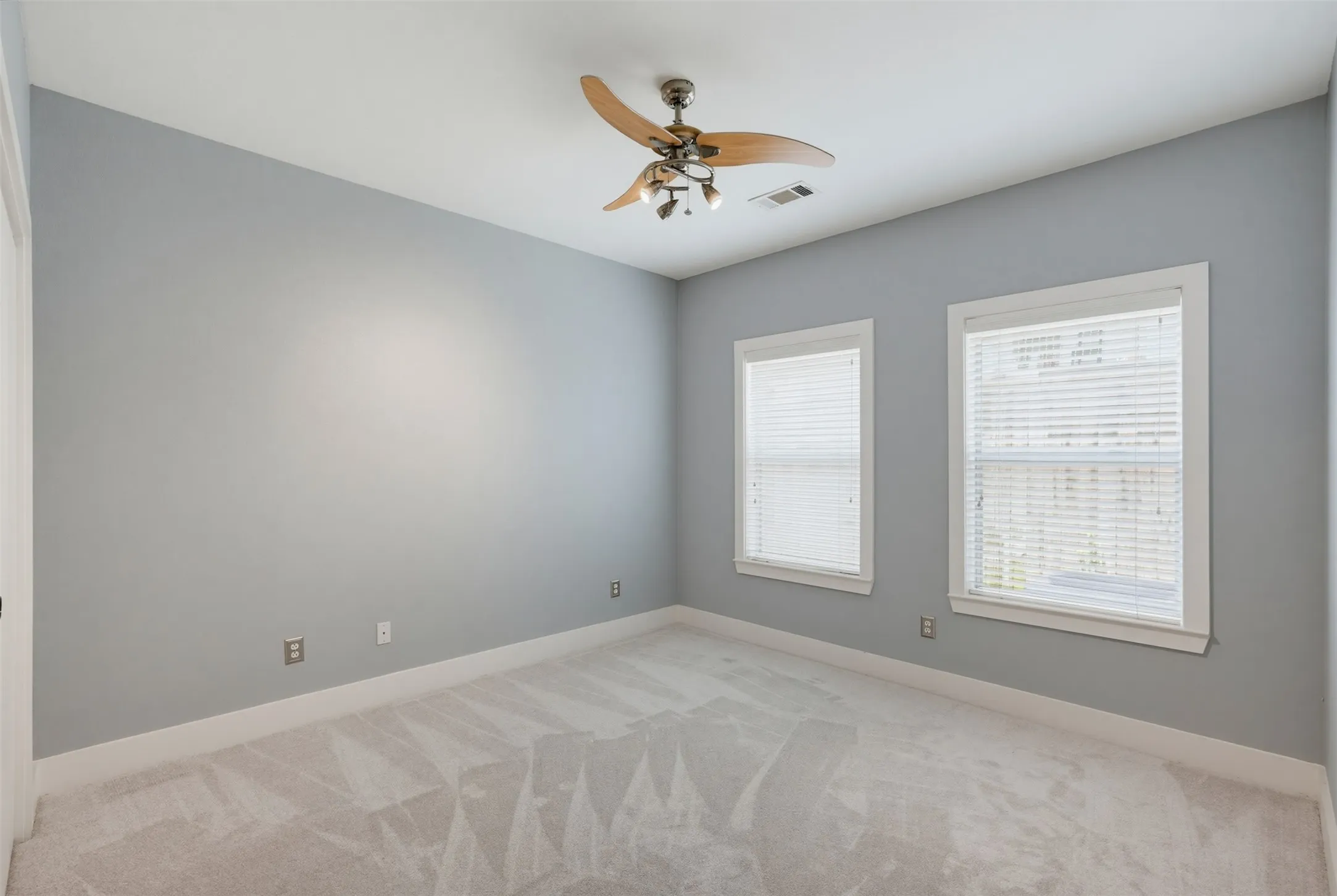 Carpeted room with light grey walls, two windows featuring white blinds, and a ceiling fan with integrated lighting