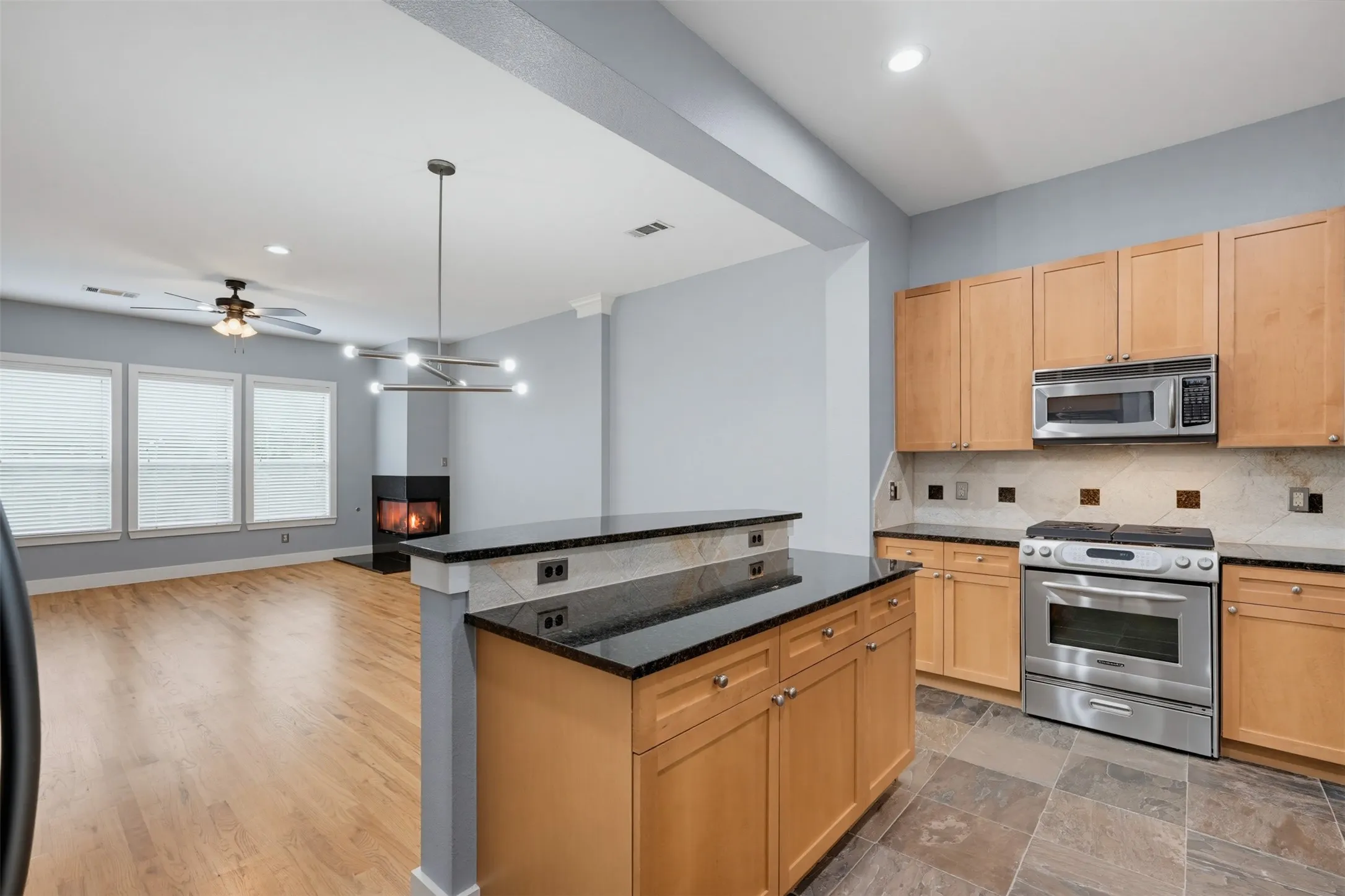 Kitchen and living area featuring a two-tiered island with a black countertop, natural wood-finish cabinetry, stainless steel appliances, and a tiled backsplash