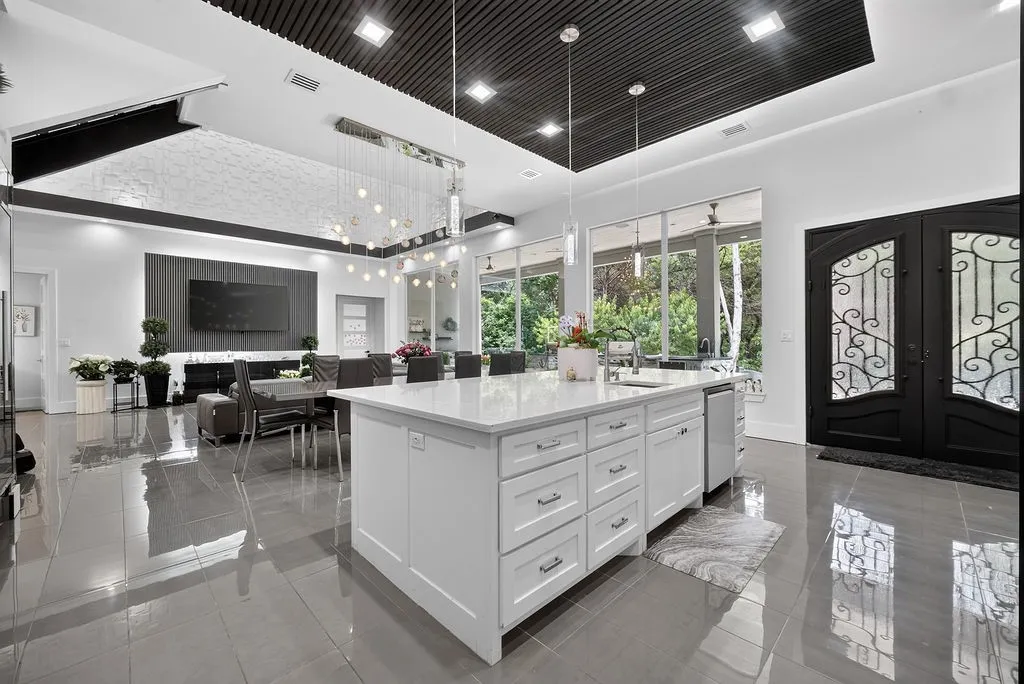 Spacious open-concept interior featuring a substantial kitchen island with a light-toned countertop, a contemporary linear chandelier, large format polished floor tiles, a decorative double entry door with wrought iron detailing, and an accent ceiling with recessed lighting
