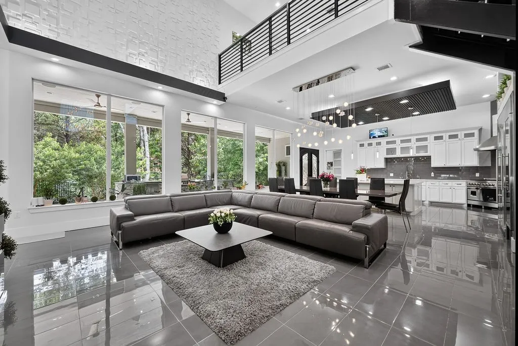 Spacious open-concept interior featuring large format tile flooring, a two-story ceiling with a balcony overlook, a modern kitchen with white cabinetry and stainless steel appliances, and extensive windows providing abundant natural light