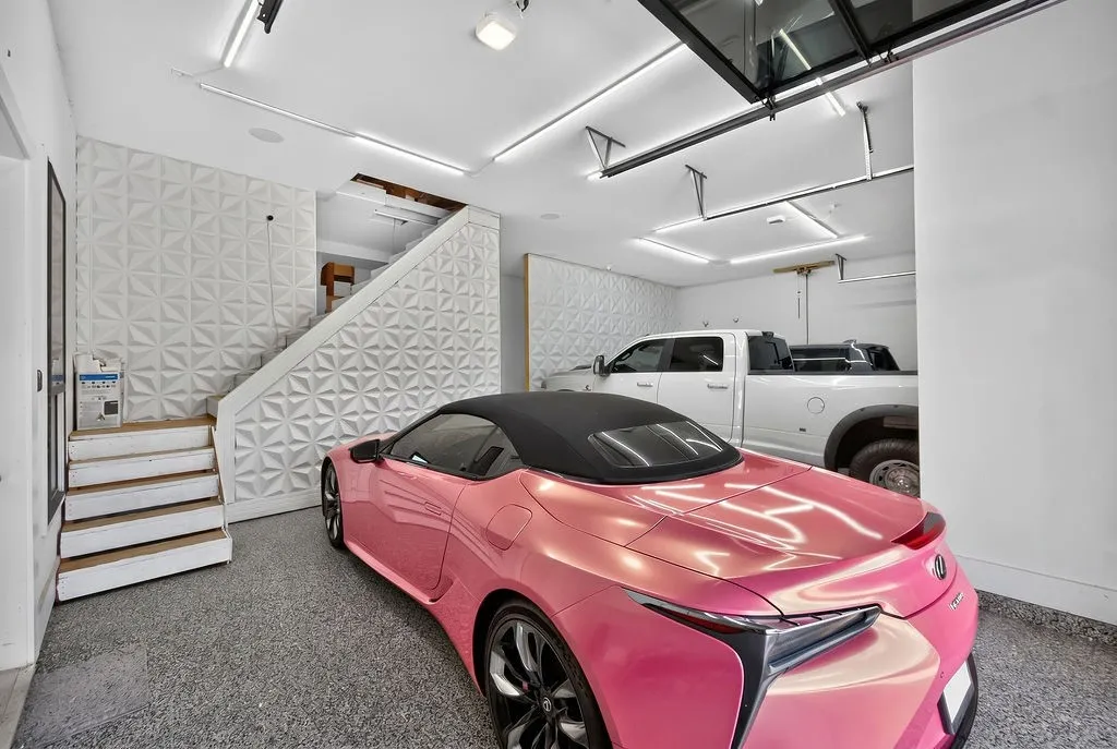 Two-car garage featuring a glass panel overhead door, epoxy flooring, linear LED lighting, and a textured accent wall with integrated staircase