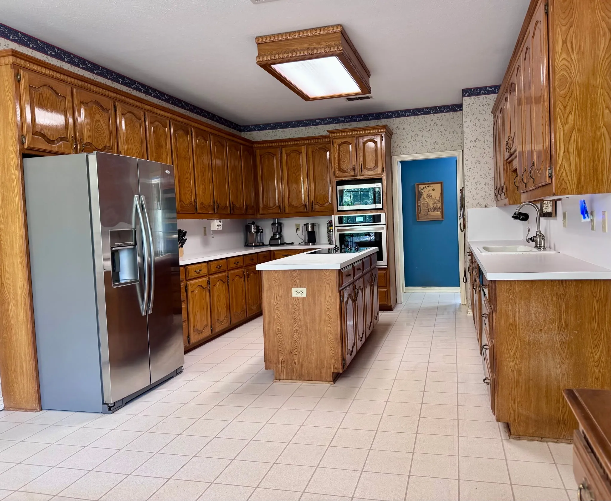 Spacious kitchen featuring extensive wood cabinetry, white countertops, a central island with integrated cooktop, stainless steel refrigerator, and built-in oven and microwave