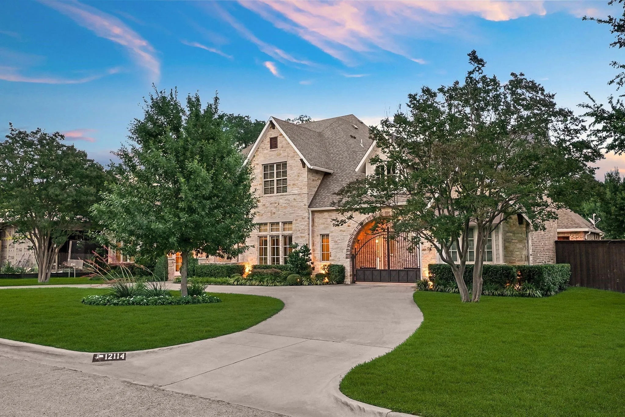Stone facade residence featuring a curved concrete driveway, mature trees, manicured lawn, arched entryway, and integrated exterior lighting