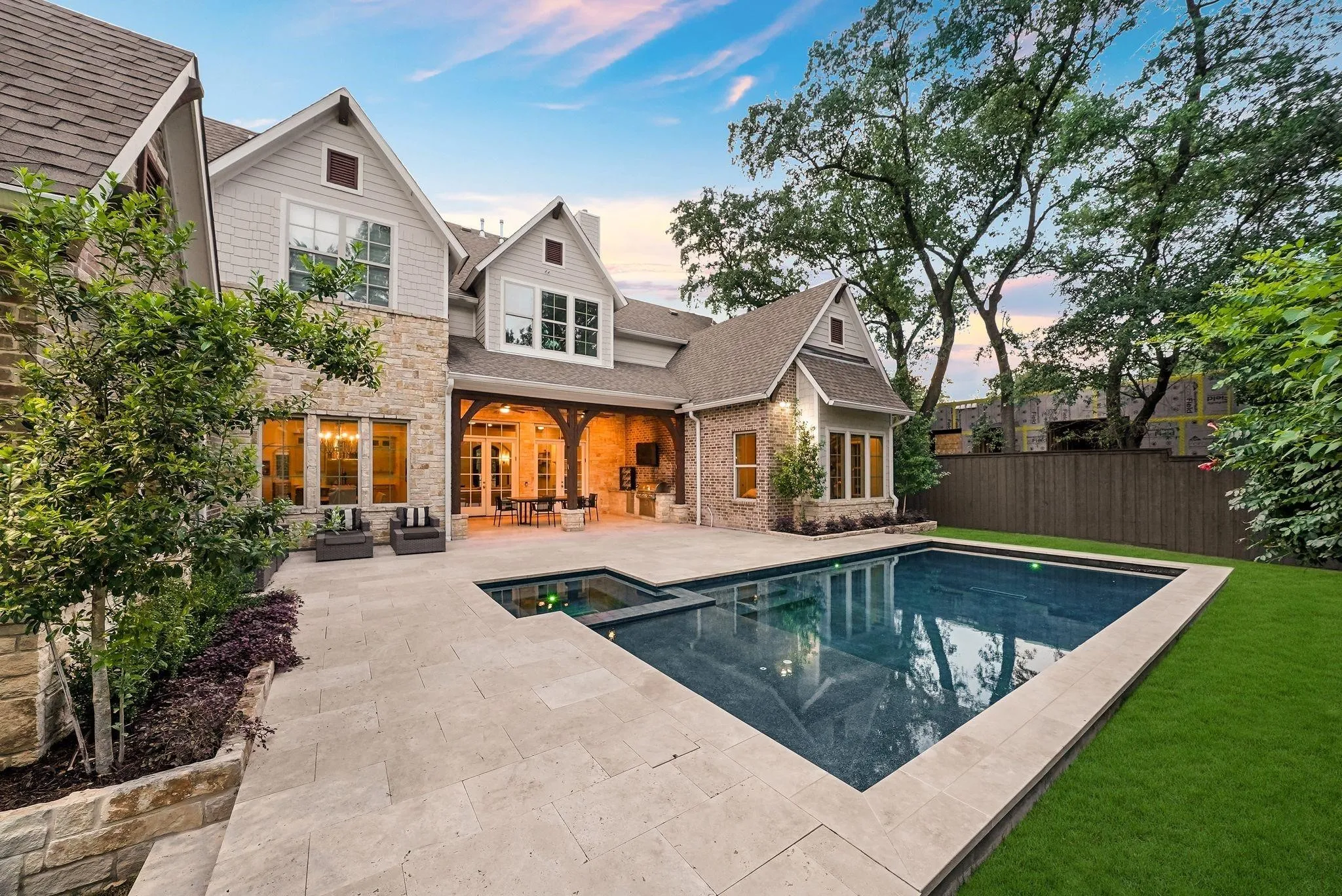 Exterior stone and siding residence featuring a swimming pool, expansive paved patio, covered outdoor living area with wood beam accents, and mature landscaping