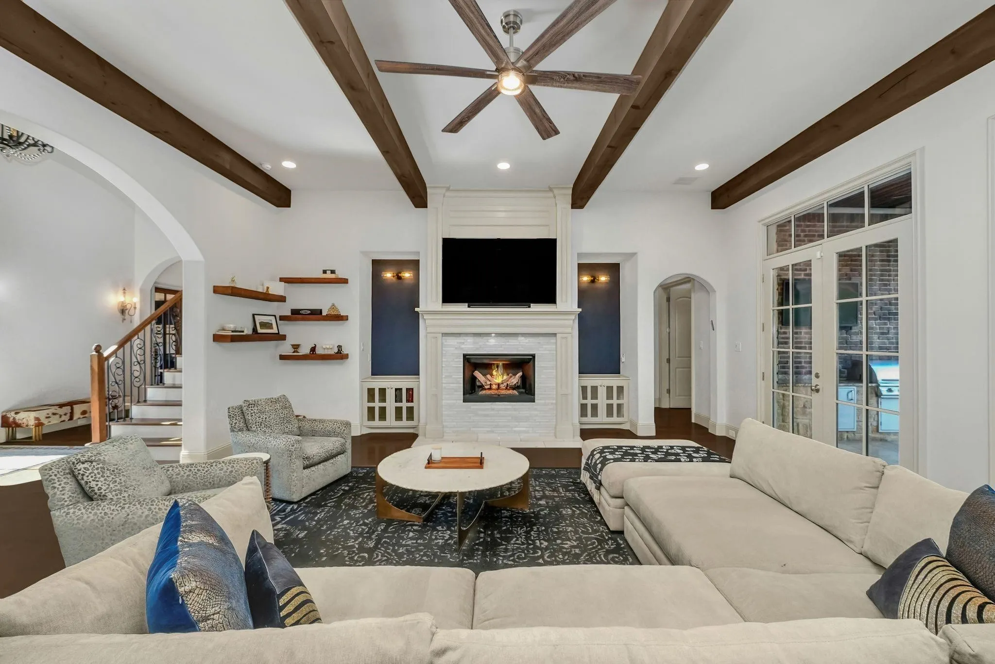 Living room featuring exposed ceiling beams, a white stacked stone fireplace with a mantel, built-in shelving, French doors, and an arched entry to the staircase