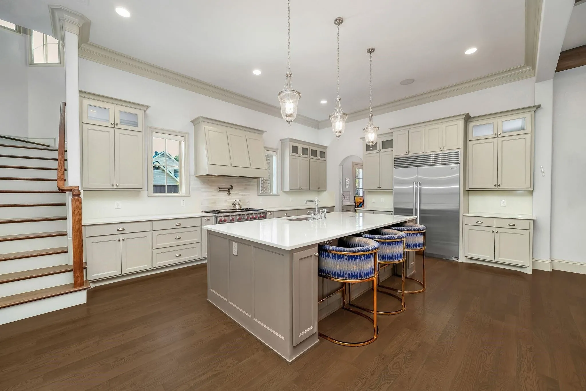Spacious kitchen featuring an expansive central island with a solid surface countertop, stainless steel appliances, extensive cabinetry with glass-front accents, dark wood-finish flooring, and pendant lighting