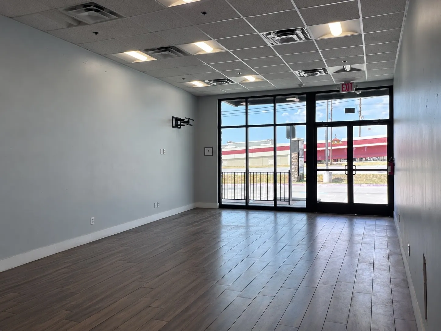 Expansive commercial space featuring wood-finish flooring, a suspended acoustic tile ceiling with integrated lighting, and large front windows with a glass entry door