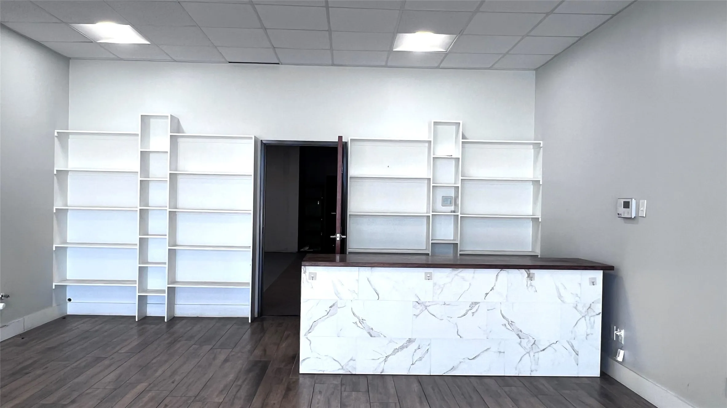 Wood-finish flooring, built-in shelving, dropped ceiling with recessed lighting, and a white and gray marbled counter with a dark wood-finish top