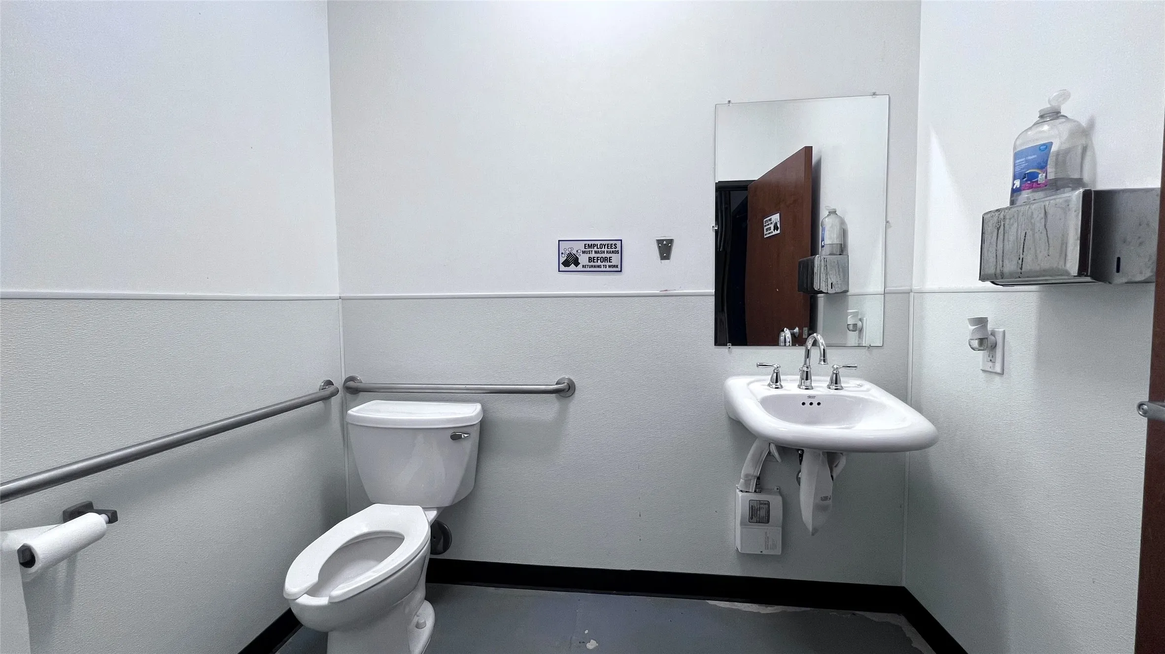 White toilet with grab bars, wall-mounted sink with mirror, and dark flooring
