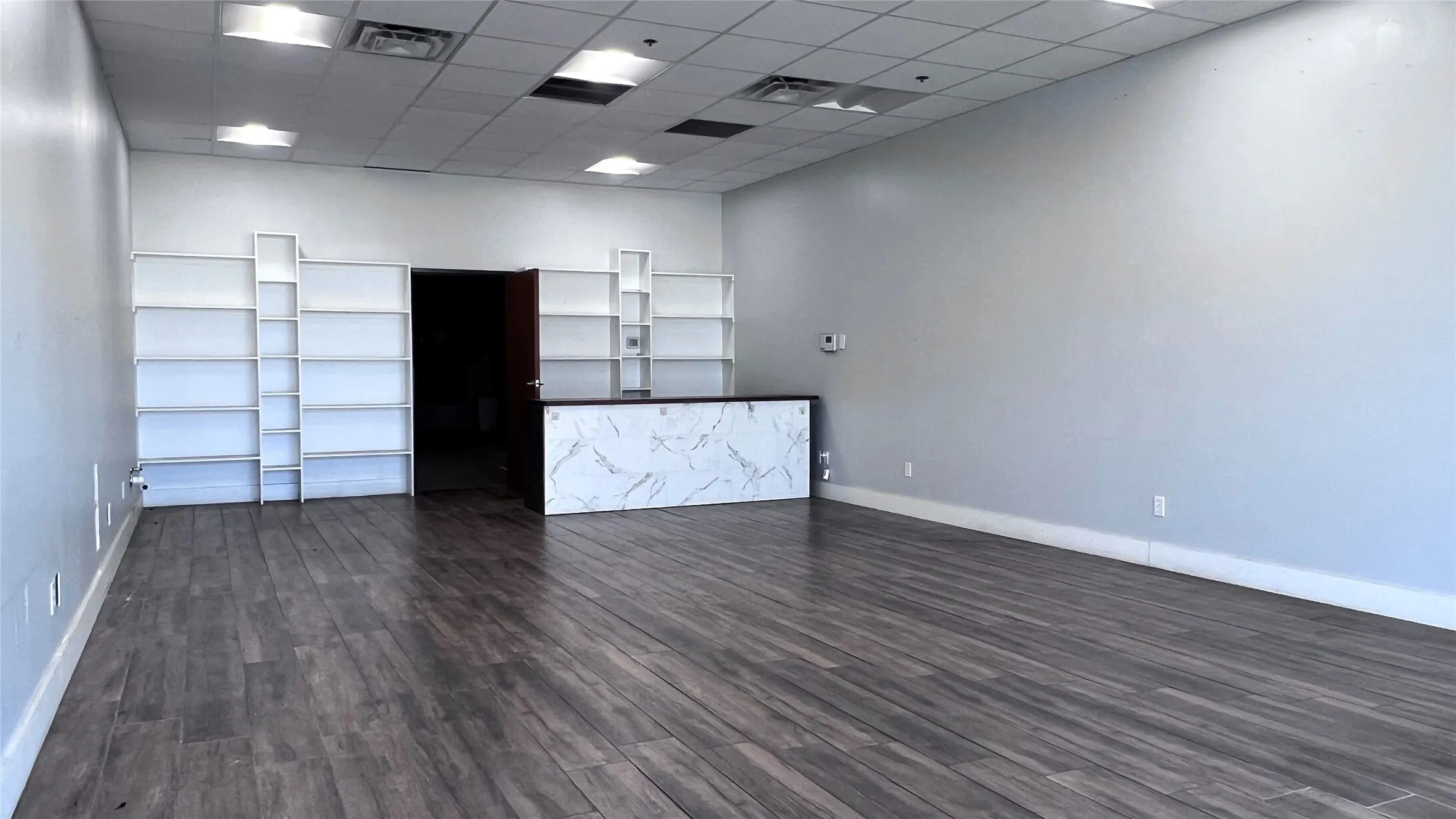 Spacious open-concept commercial interior featuring wood-finish flooring, recessed lighting, built-in shelving, and a marble-finish reception counter