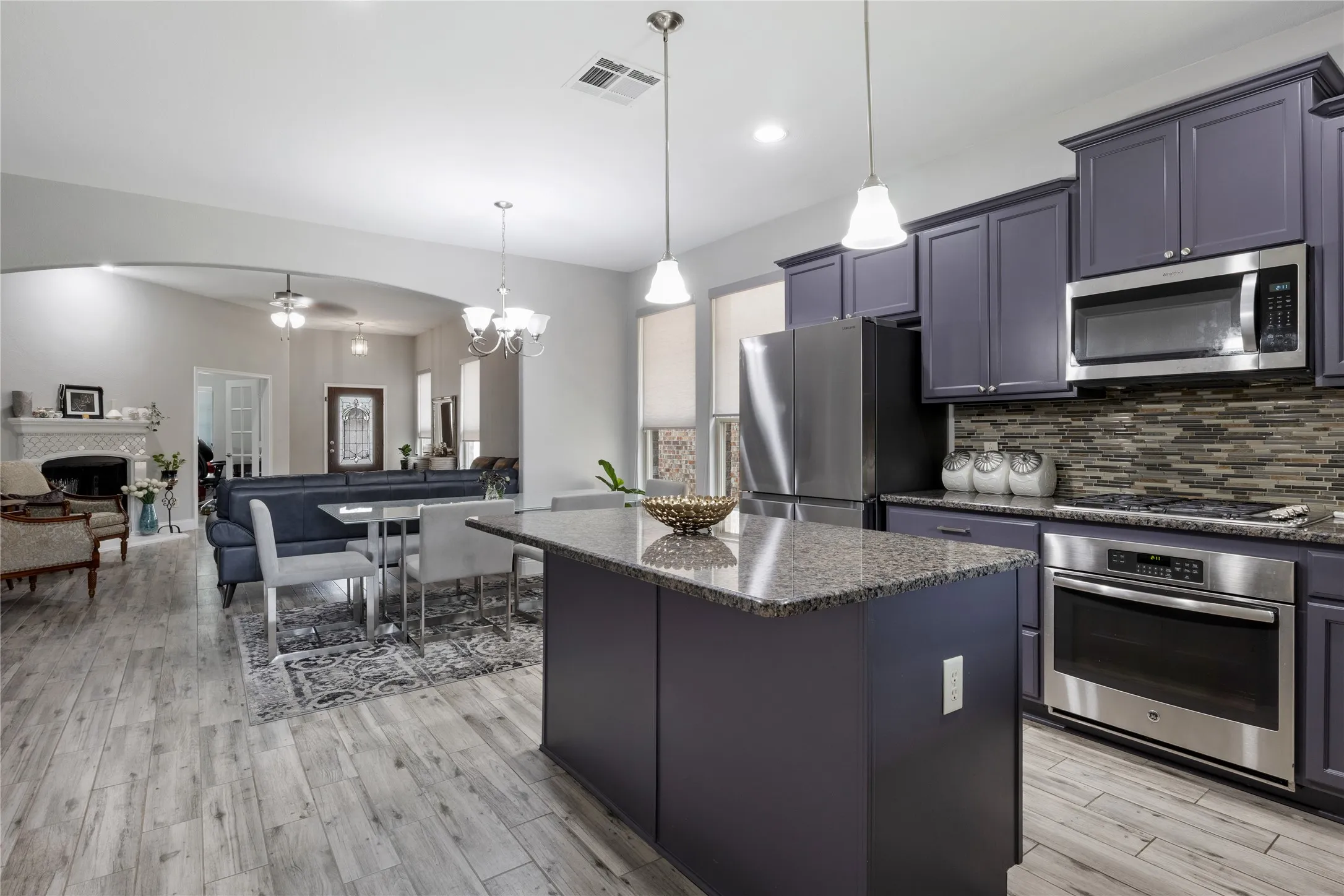 Gourmet kitchen featuring a central island with granite countertops, stainless steel appliances, a mosaic tile backsplash, custom cabinetry, and pendant lighting, opening to an expansive living area with arched entryways and wood-finish flooring