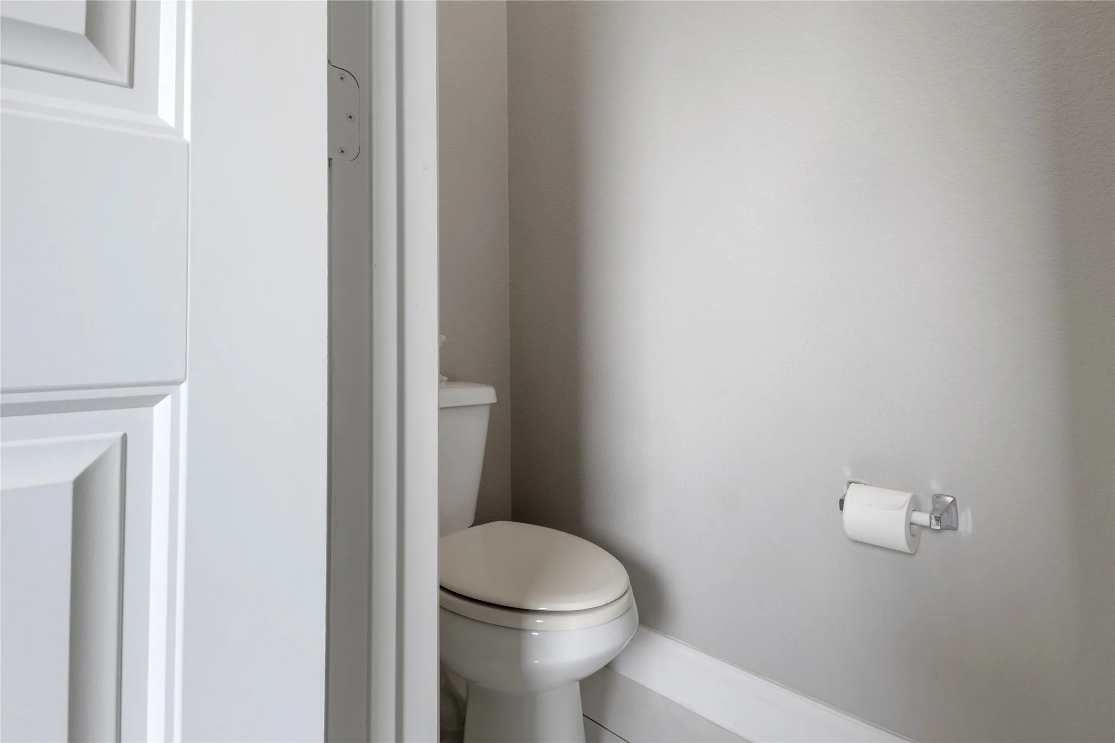 White paneled door opening to a water closet featuring a porcelain toilet with a beige seat, light gray walls, and white baseboards