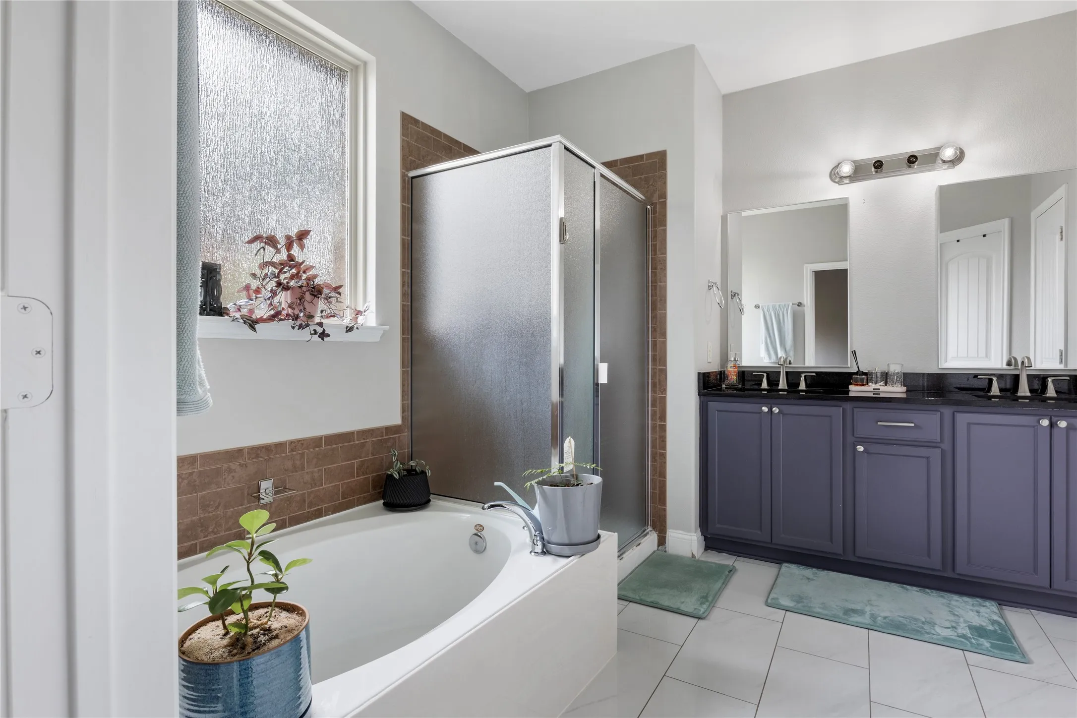 Bathroom featuring a built-in soaking tub with surrounding tile, a separate glass-enclosed shower with tiled walls, and a double vanity with dark cabinetry, dark countertop, and dual sinks