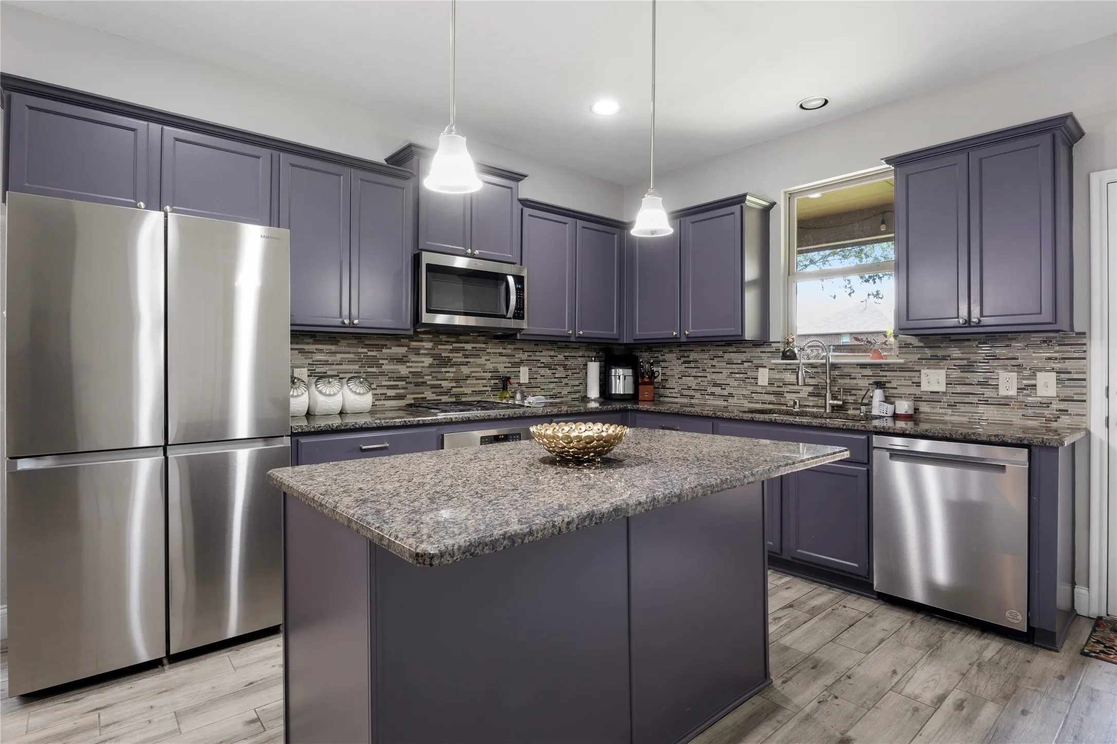 Kitchen featuring a central island with a stone-finish countertop, wood-finish flooring, purple cabinetry, stainless steel appliances, and a mosaic tile backsplash