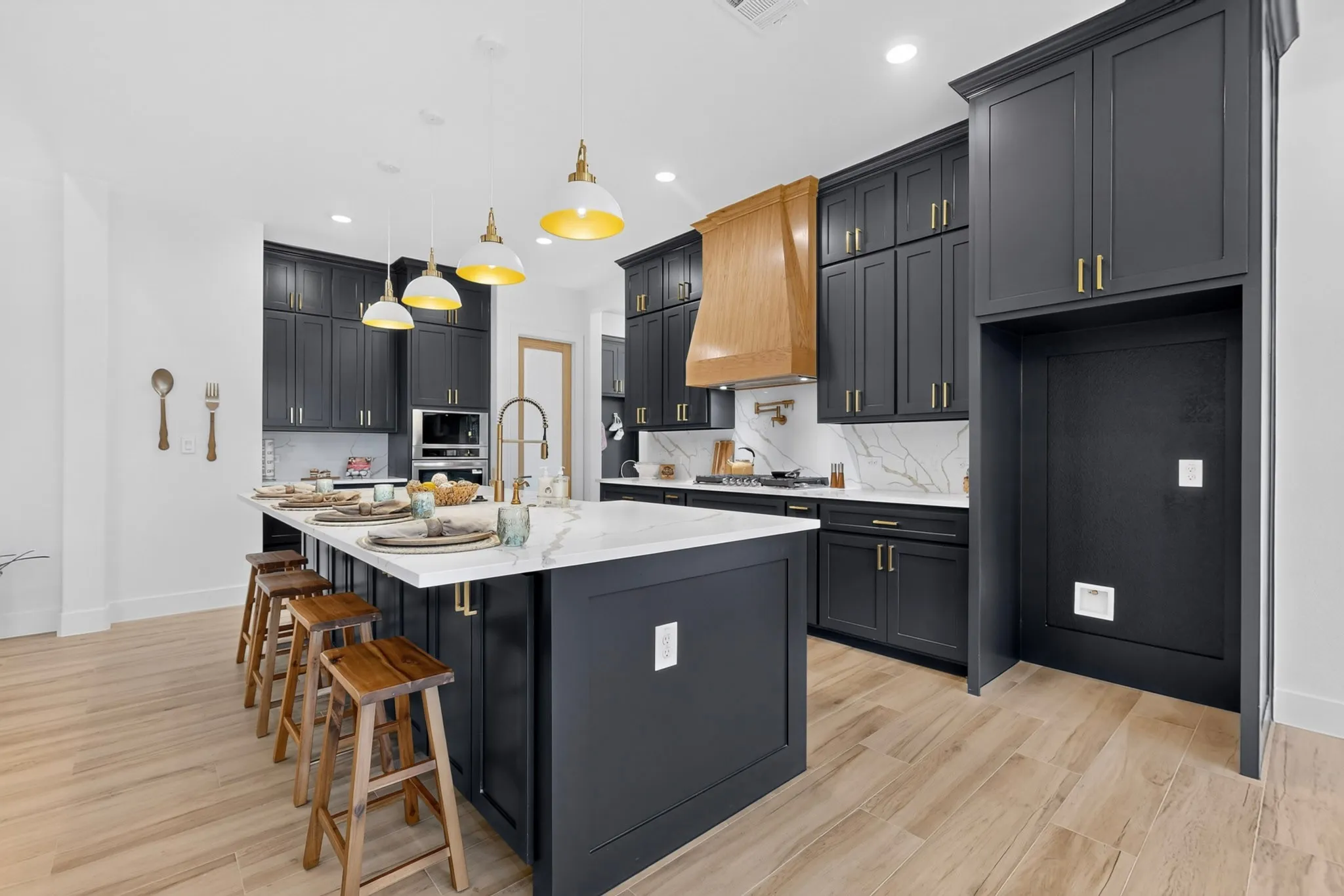 Modern kitchen featuring dark cabinetry with gold-tone hardware, a large central island with seating, wood-finish flooring, a wood range hood, and pendant lighting