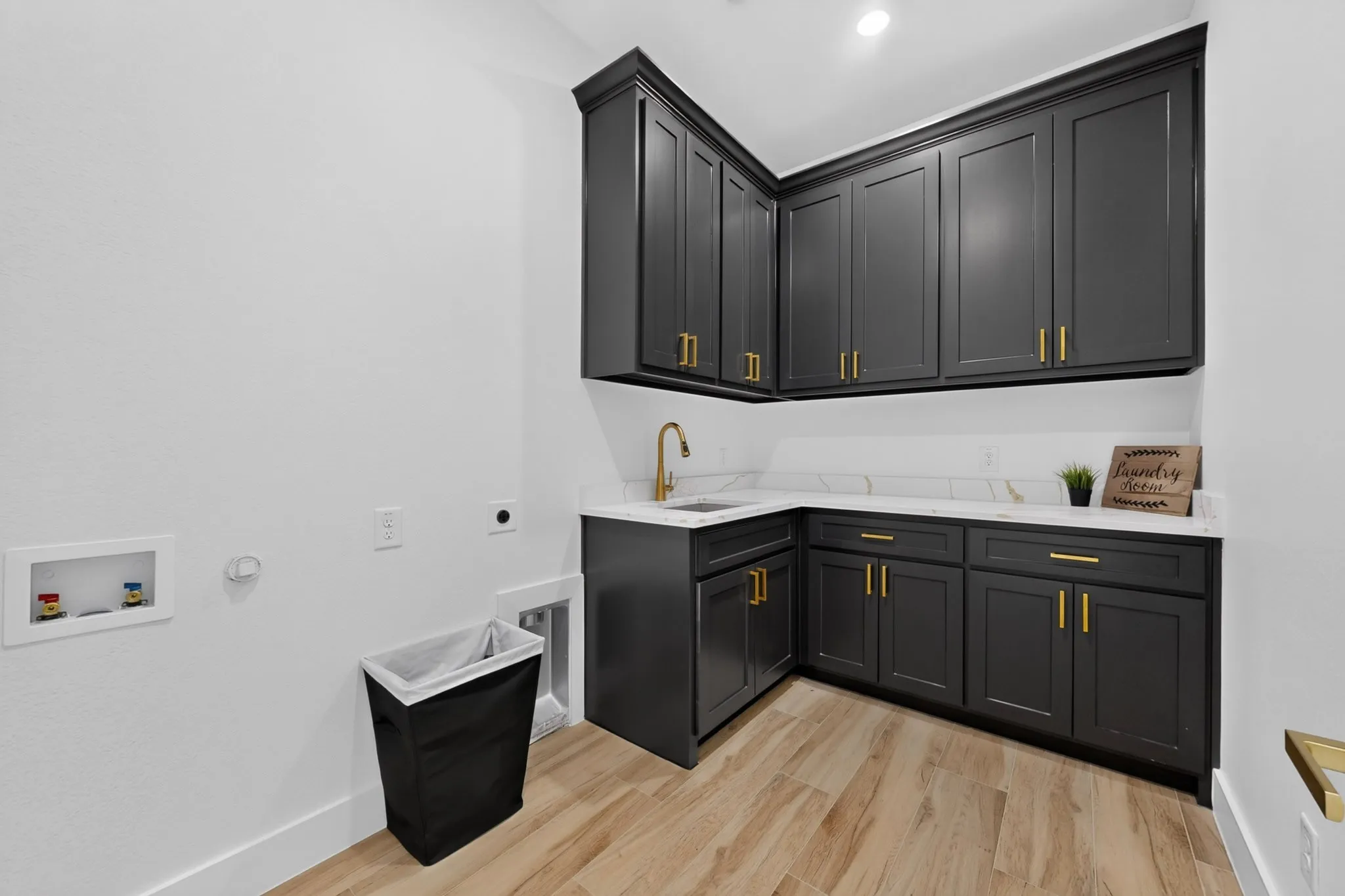 Laundry area featuring dark cabinetry with gold-tone hardware, white countertops, a single-basin sink with a gold-tone faucet, and wood-finish flooring