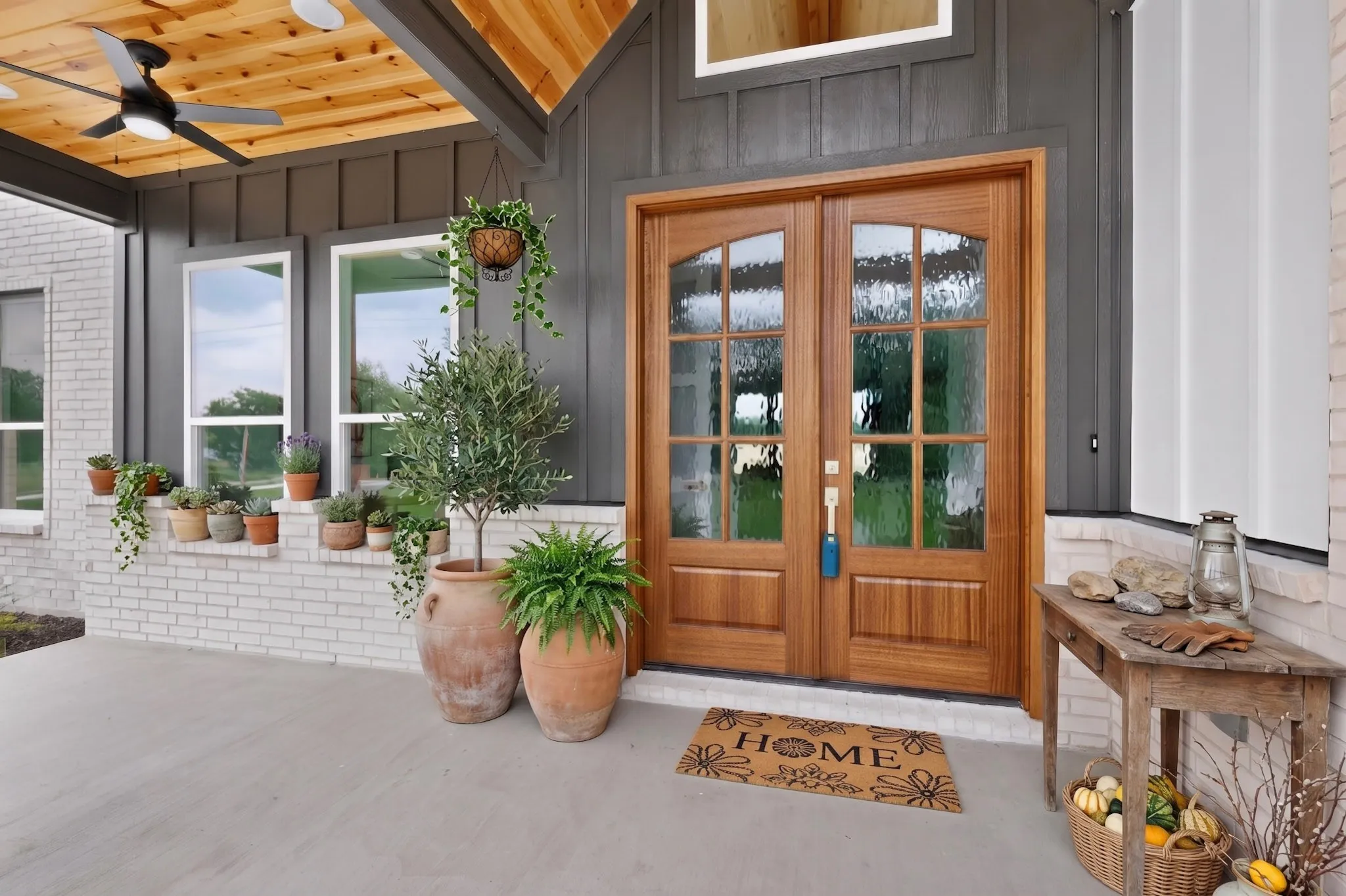 Inviting front porch featuring double wood-finish entry doors with arched glass inserts, a wood-plank ceiling with a ceiling fan, and board and batten siding details