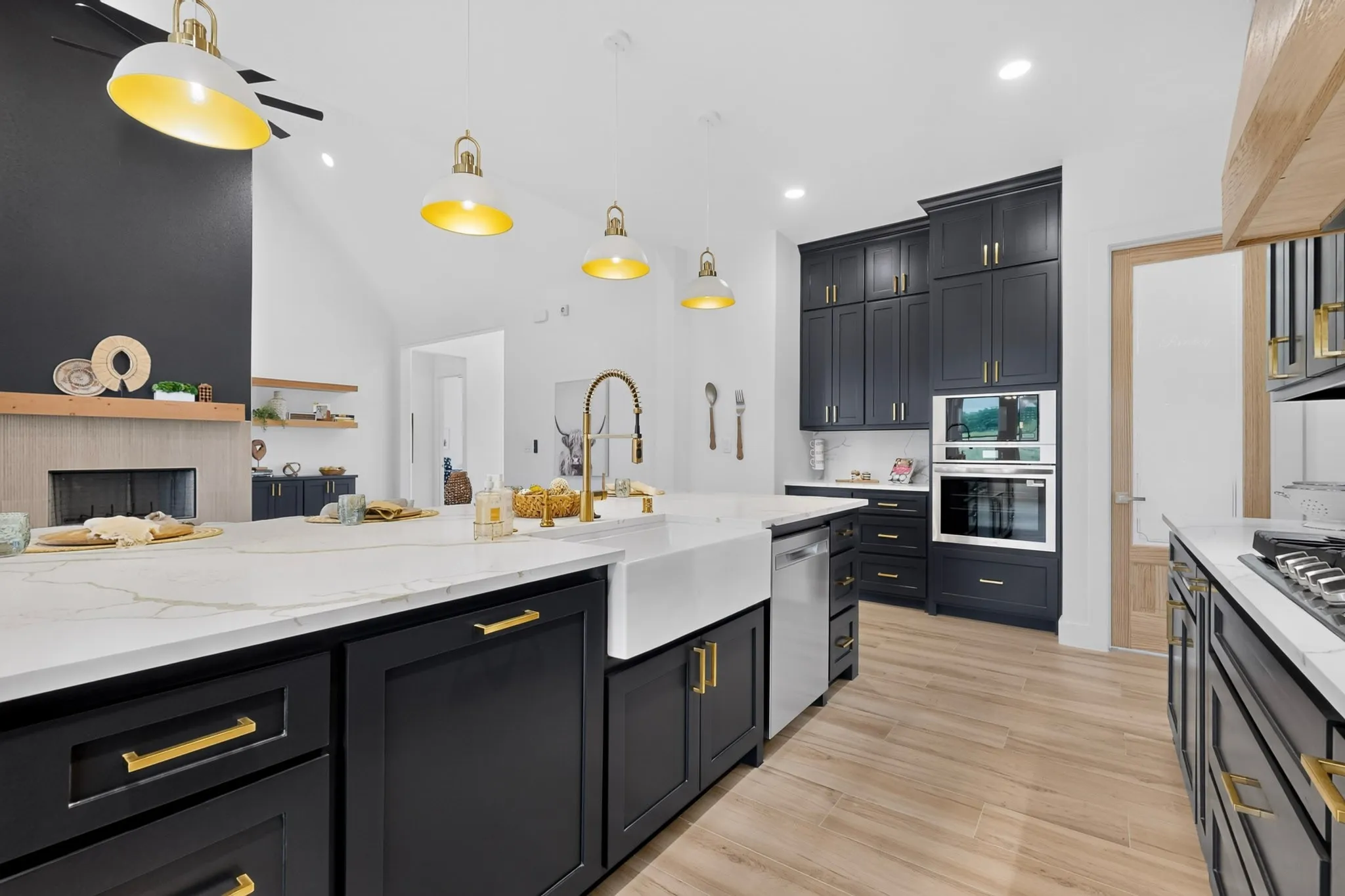 Spacious kitchen featuring a large central island with a white farmhouse sink, dark cabinetry with gold hardware, stainless steel appliances, and light wood-finish flooring