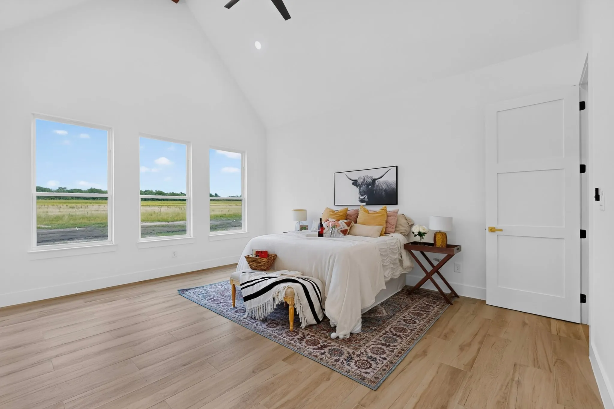 Vaulted ceiling with an exposed beam, light wood-finish flooring, three large windows, three-panel interior door, and a ceiling fan