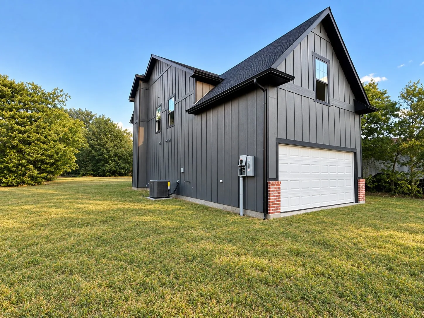 Board and batten siding, dark shingle roofing, white garage door, brick accents, and an expansive lawn
