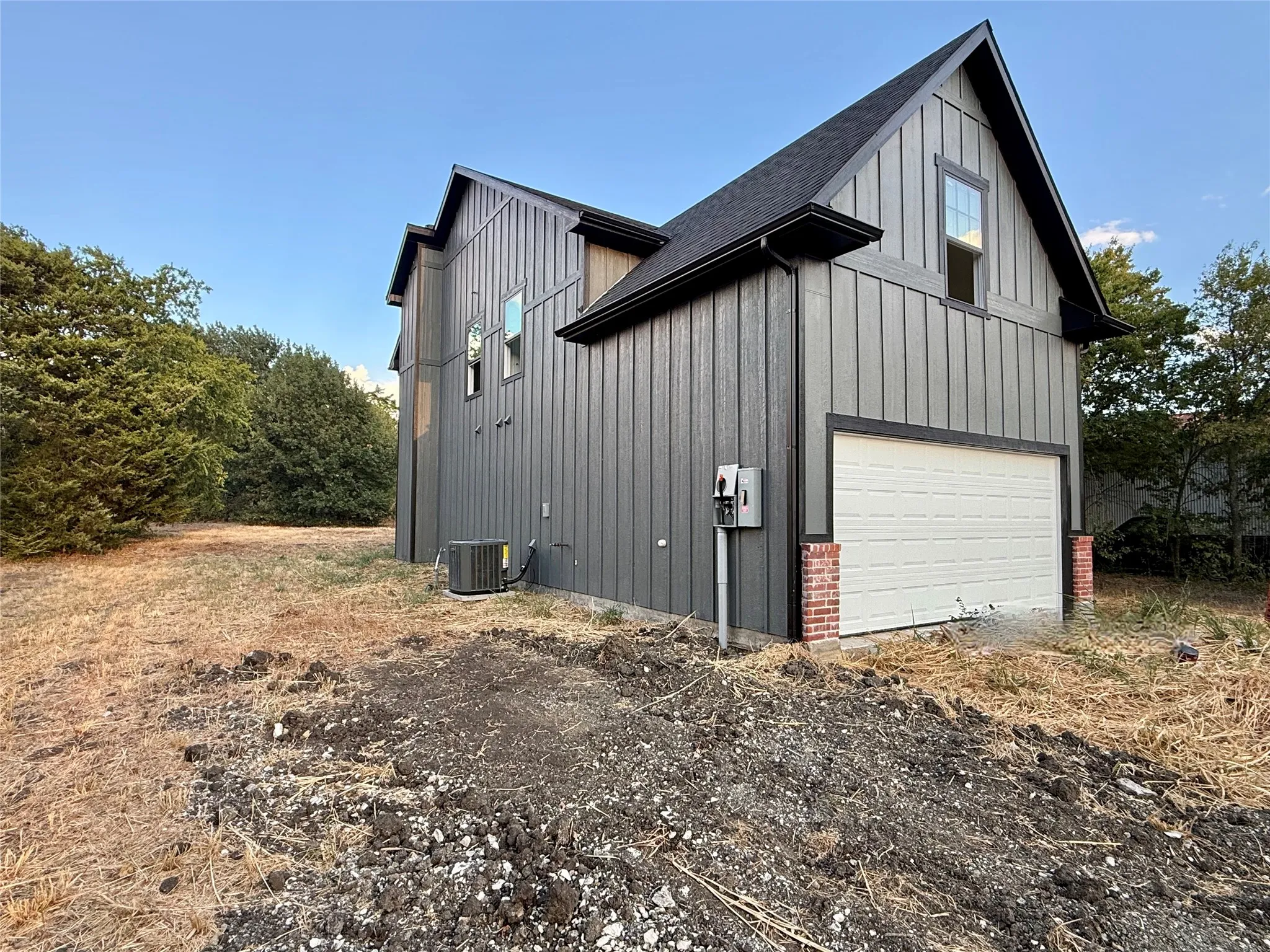 Board and batten siding, dark trim accents, attached garage with brick pilasters, upper-level window, and a dark shingle roof