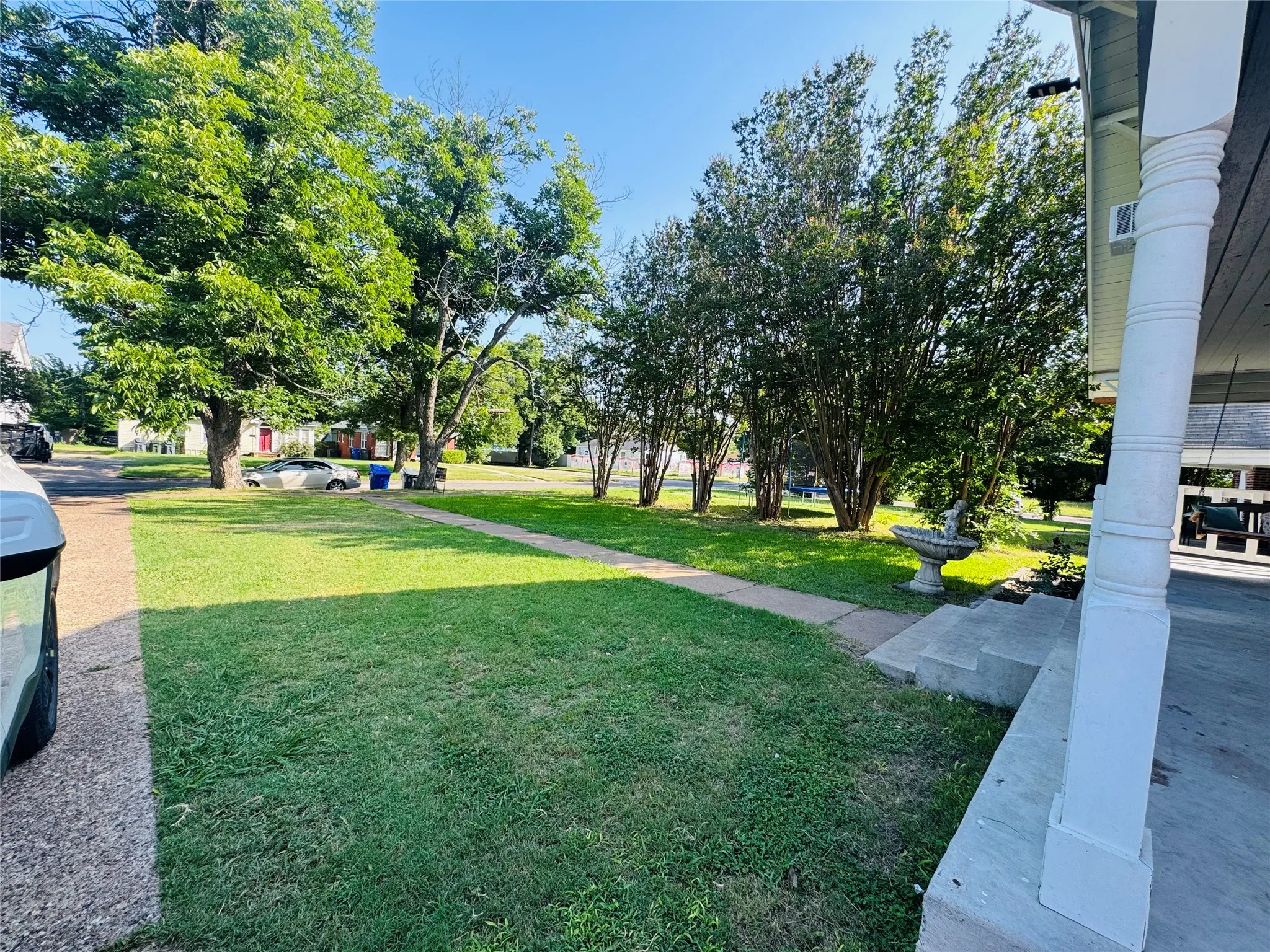 Front yard featuring a concrete walkway, mature trees, established shrubbery, and a decorative garden fountain