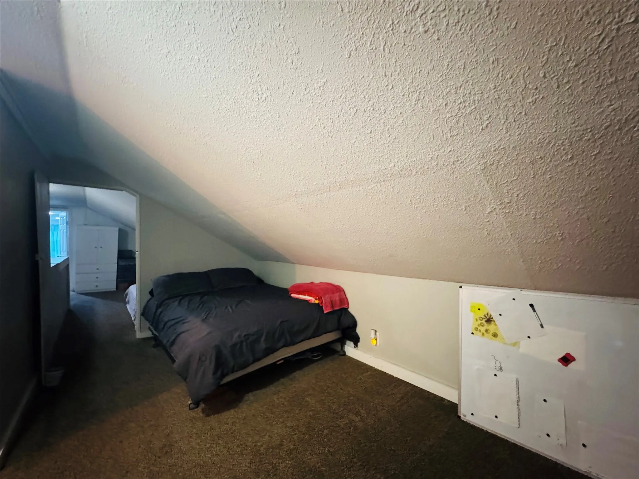 Carpeted attic space featuring a sloped ceiling with a textured finish and an open doorway leading to another area