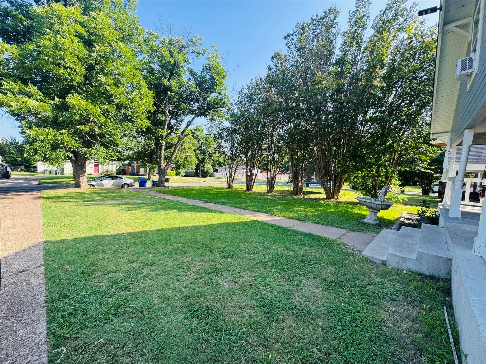 Expansive front yard featuring a concrete walkway, mature trees, a manicured lawn, and a decorative garden accent