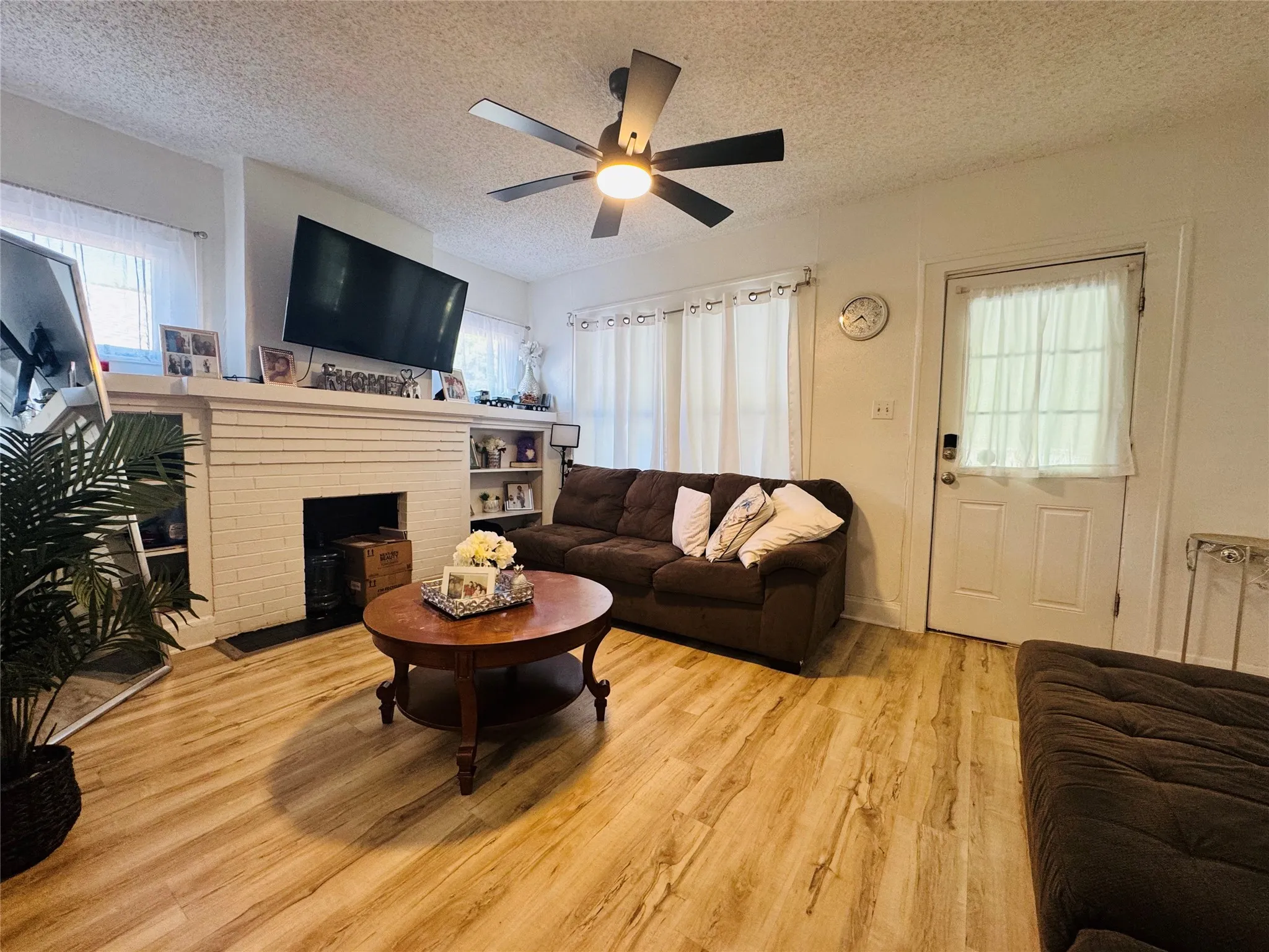 Spacious living area featuring wood-finish flooring, a white brick fireplace with mantel, built-in shelving, a ceiling fan with integrated lighting, and multiple windows