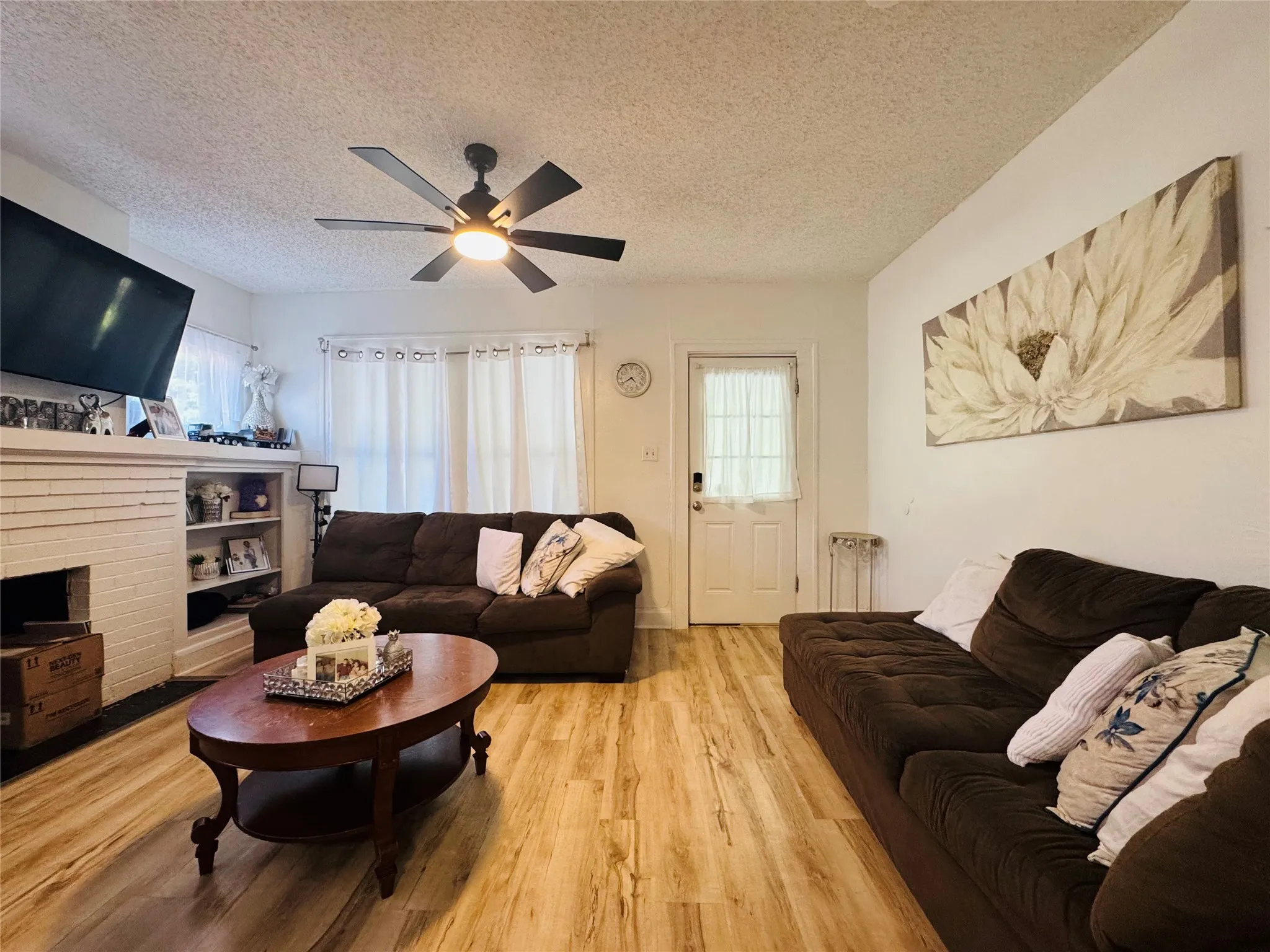 Living space featuring wood-finish flooring, a white brick fireplace with built-in shelving, a ceiling fan with integrated lighting, and white walls