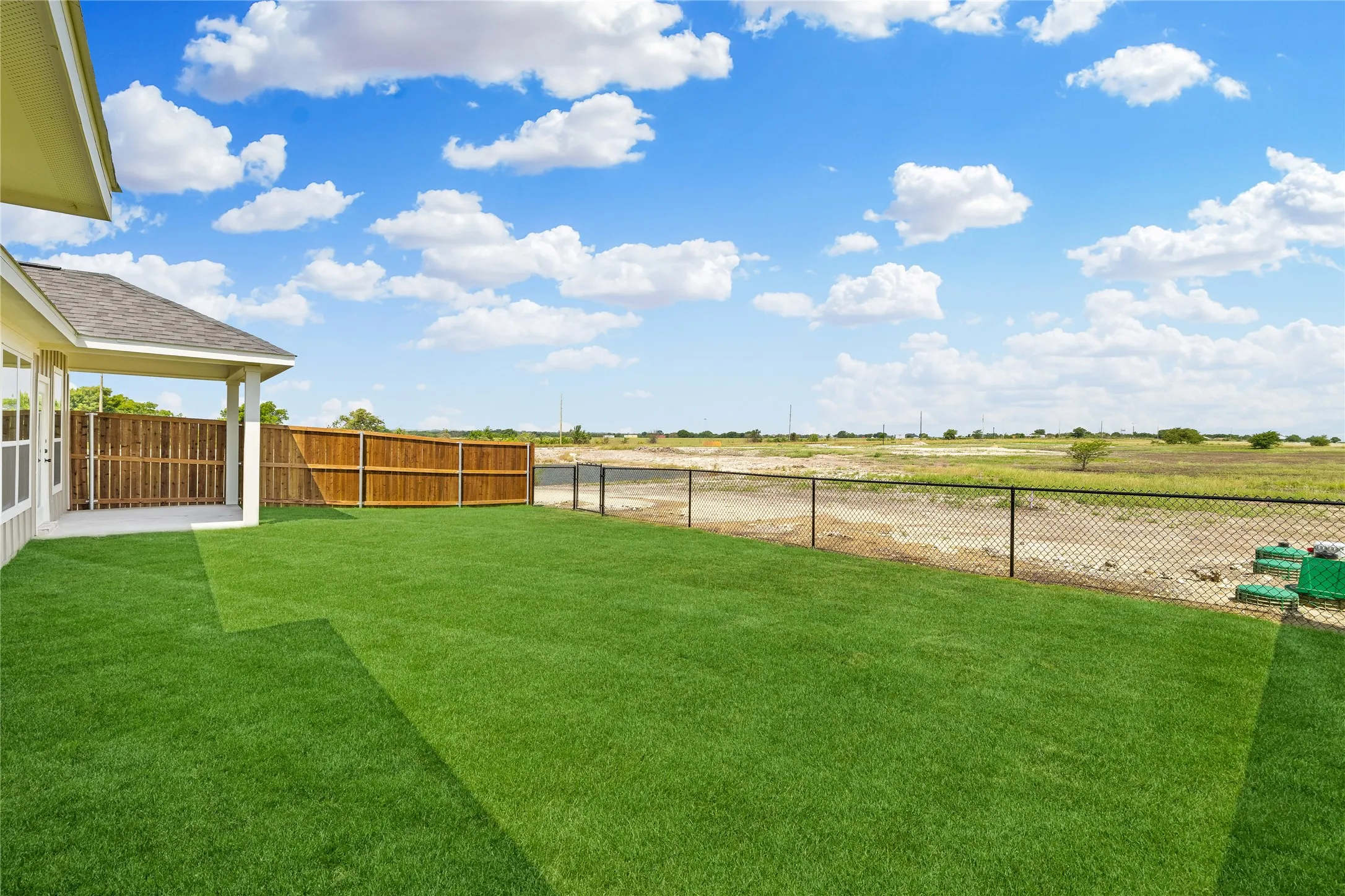 Expansive green lawn with a covered patio, wood privacy fencing, and chain-link fencing bordering open land