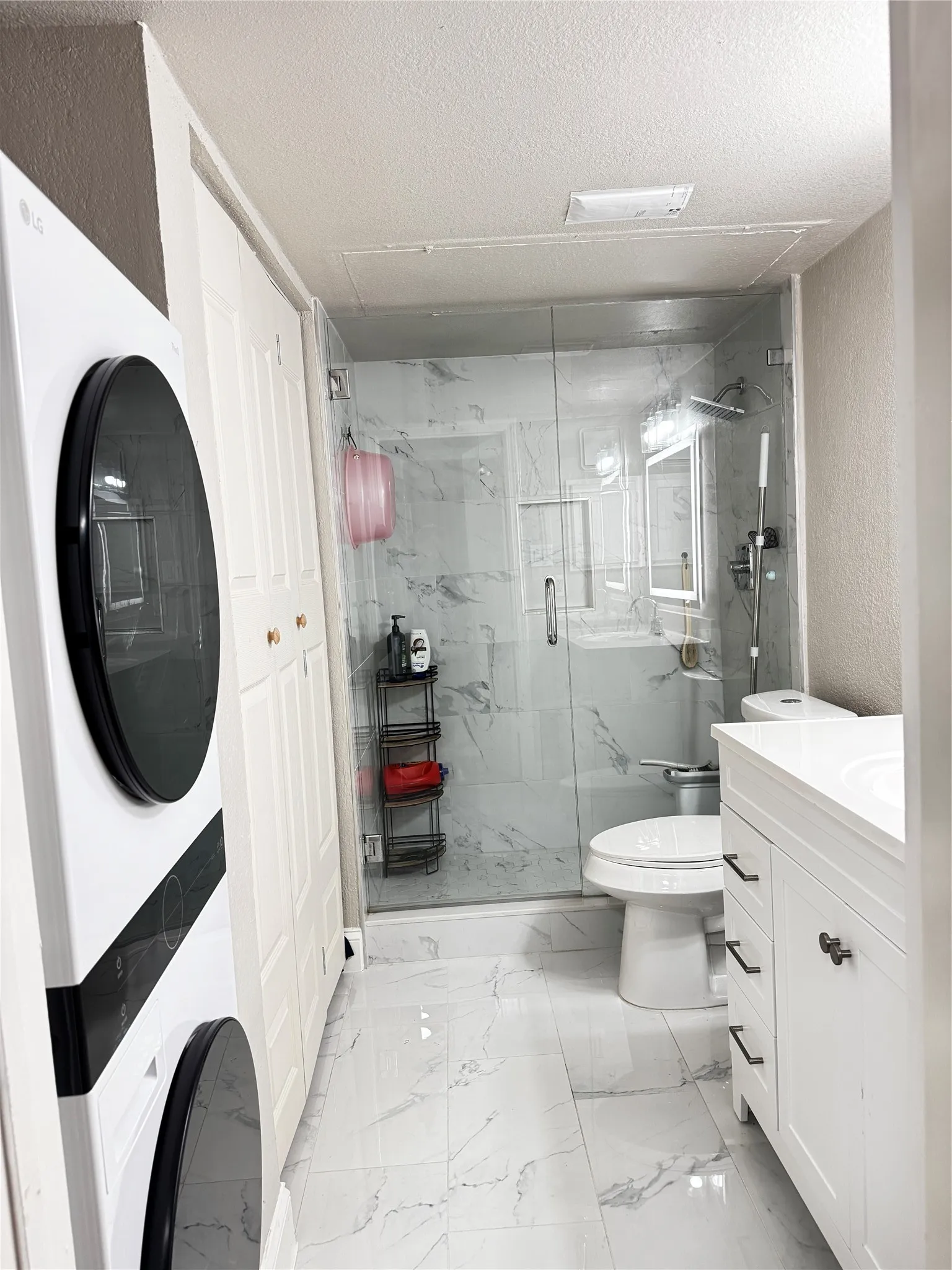 Bathroom featuring a glass-enclosed shower with marble-look wall tile, a white vanity with integrated sink and black hardware, marble-look floor tile, and a stacked washer and dryer unit