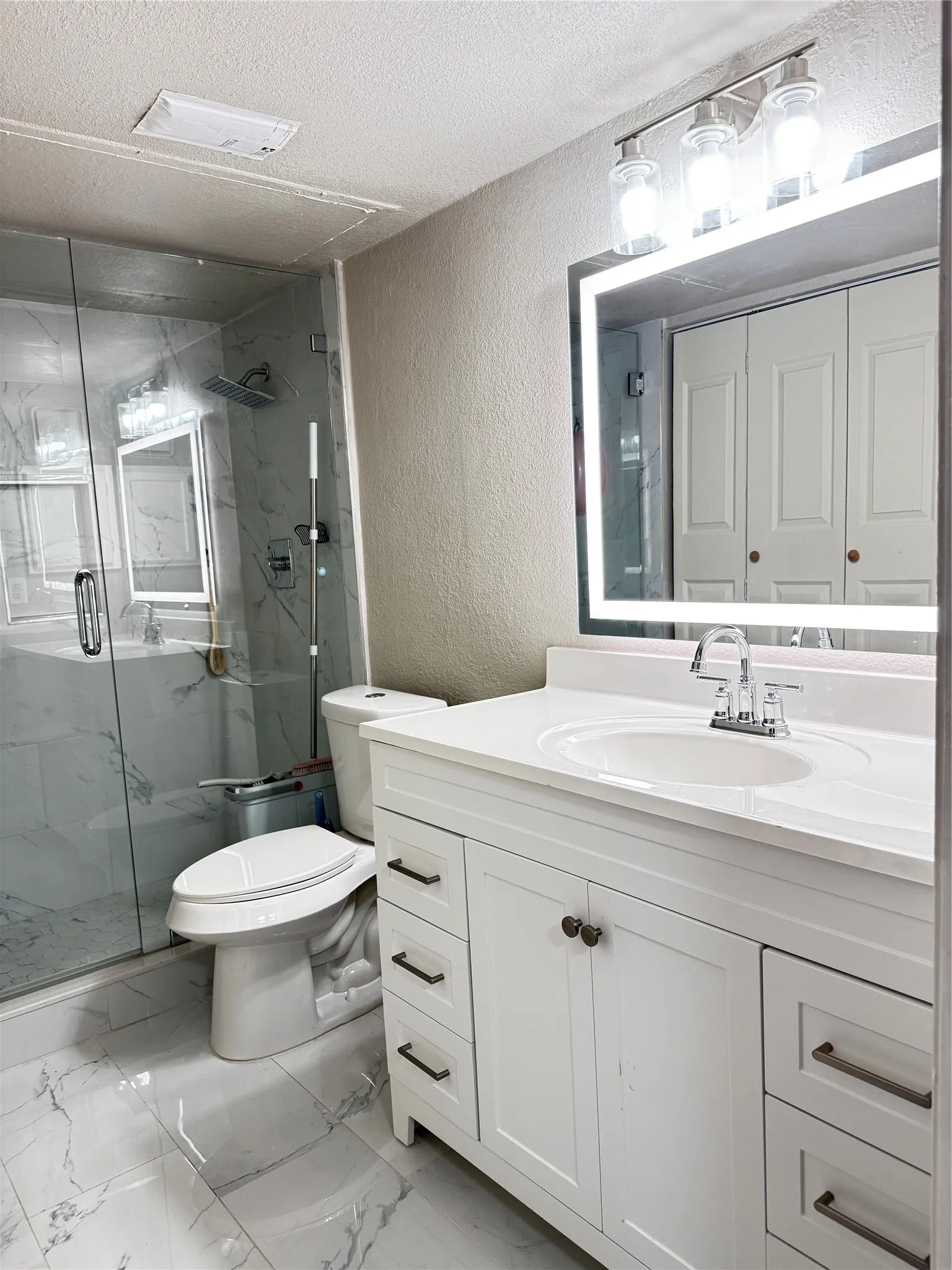 Contemporary bathroom featuring a frameless glass shower enclosure, a full vanity with a white countertop and integrated sink, a large backlit mirror, and white-finish cabinetry with dark hardware