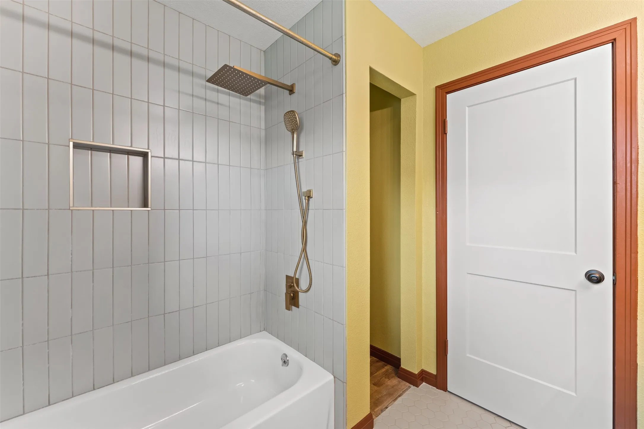 Bathtub shower with vertical subway tile surround, integrated shower niche, and gold-finish fixtures including a rain showerhead and handheld sprayer