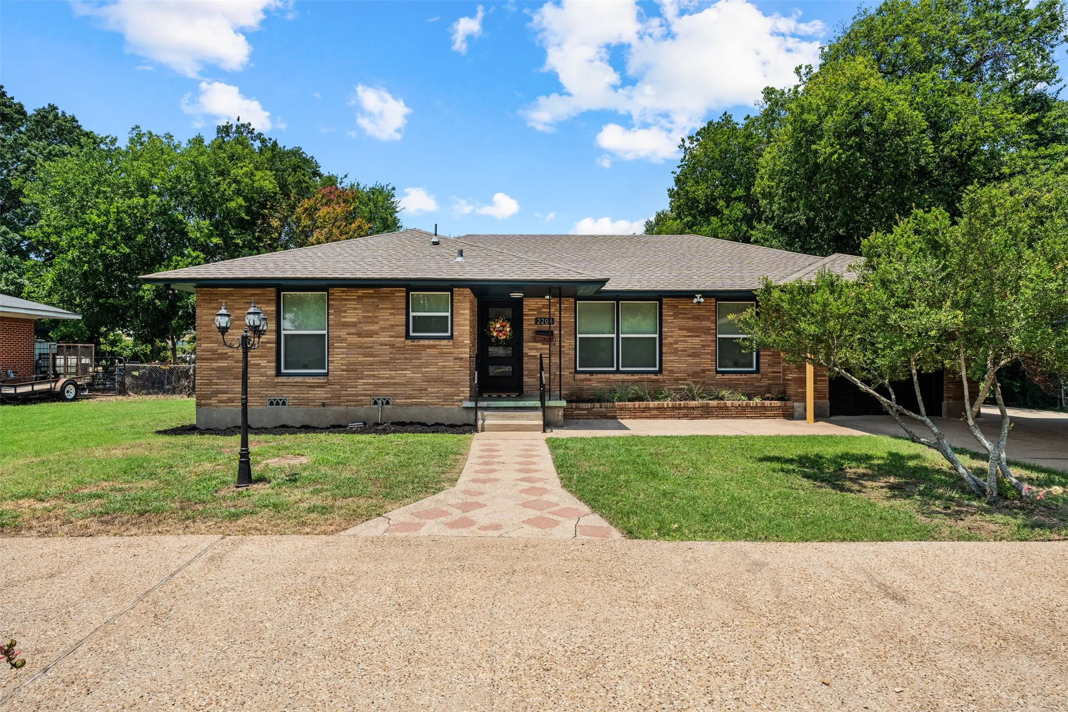 Brick facade residence featuring a patterned paver walkway, mature landscaping, a covered entry, and a multi-pane front door