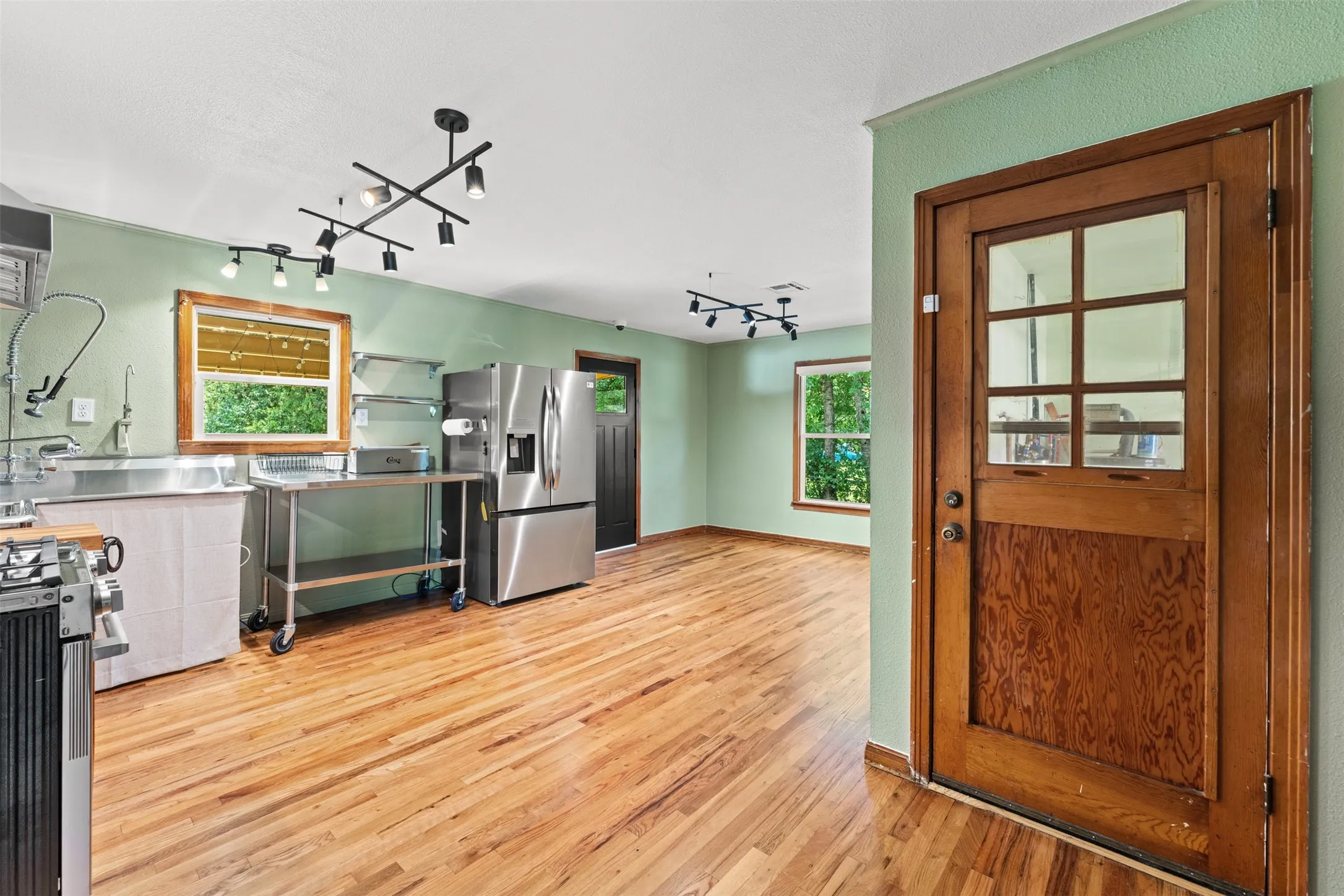 Expansive room featuring wood-finish flooring, stainless steel appliances, track lighting, and a paneled wooden door with glass inserts