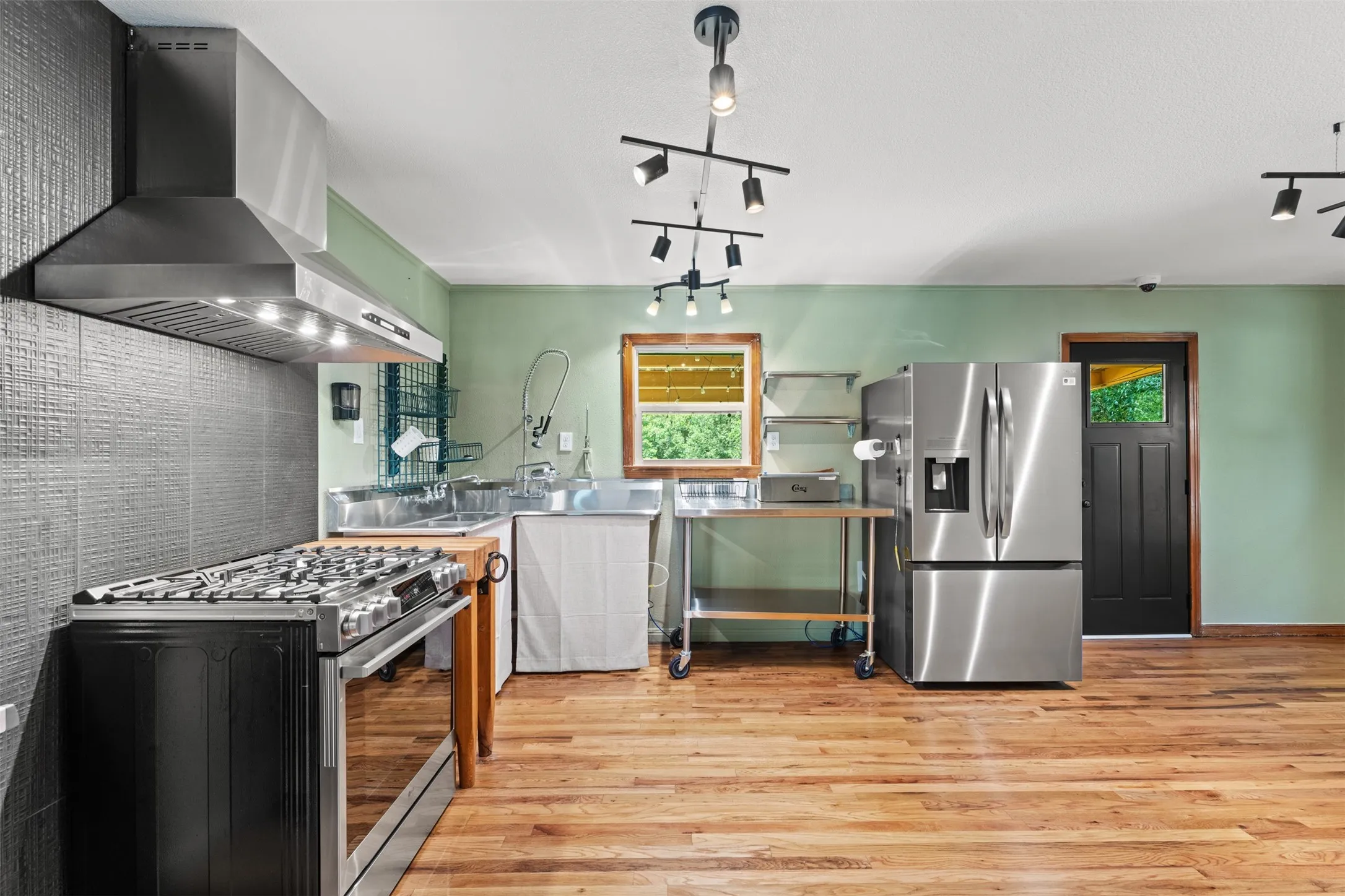 Kitchen featuring stainless steel appliances, wood-finish flooring, a commercial-style range with a substantial range hood, and a dark wood door with glass inserts