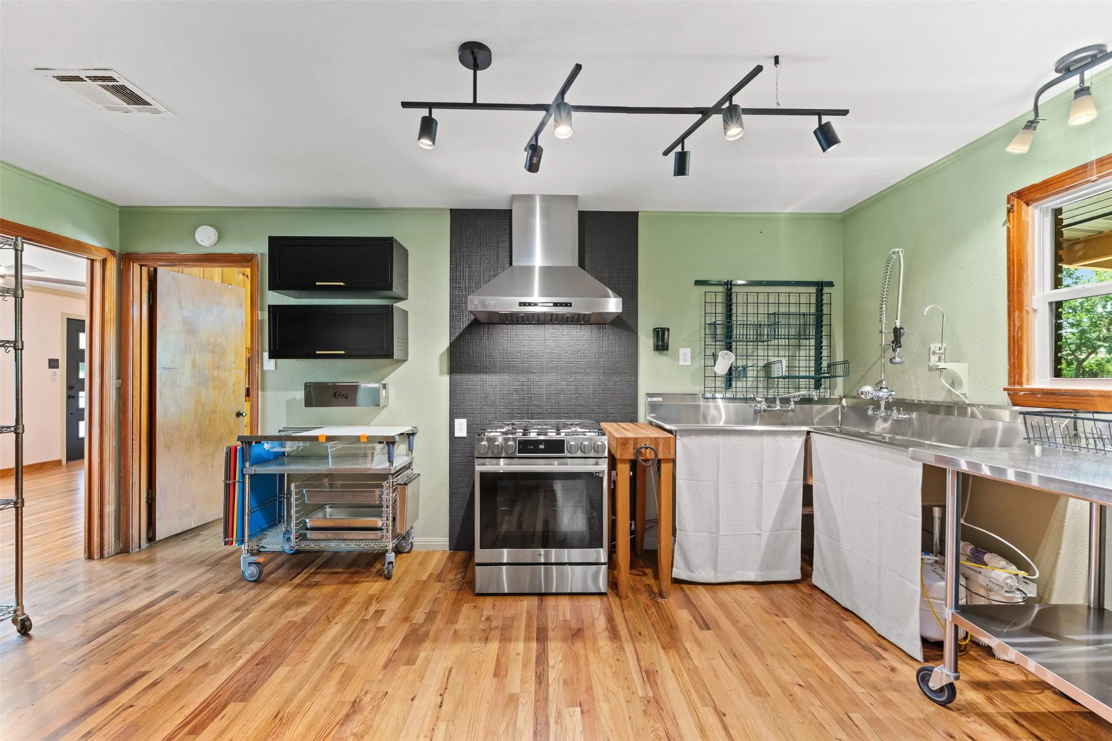 Kitchen featuring wood-finish flooring, stainless steel appliances, black cabinetry, a stainless steel range hood, and track lighting