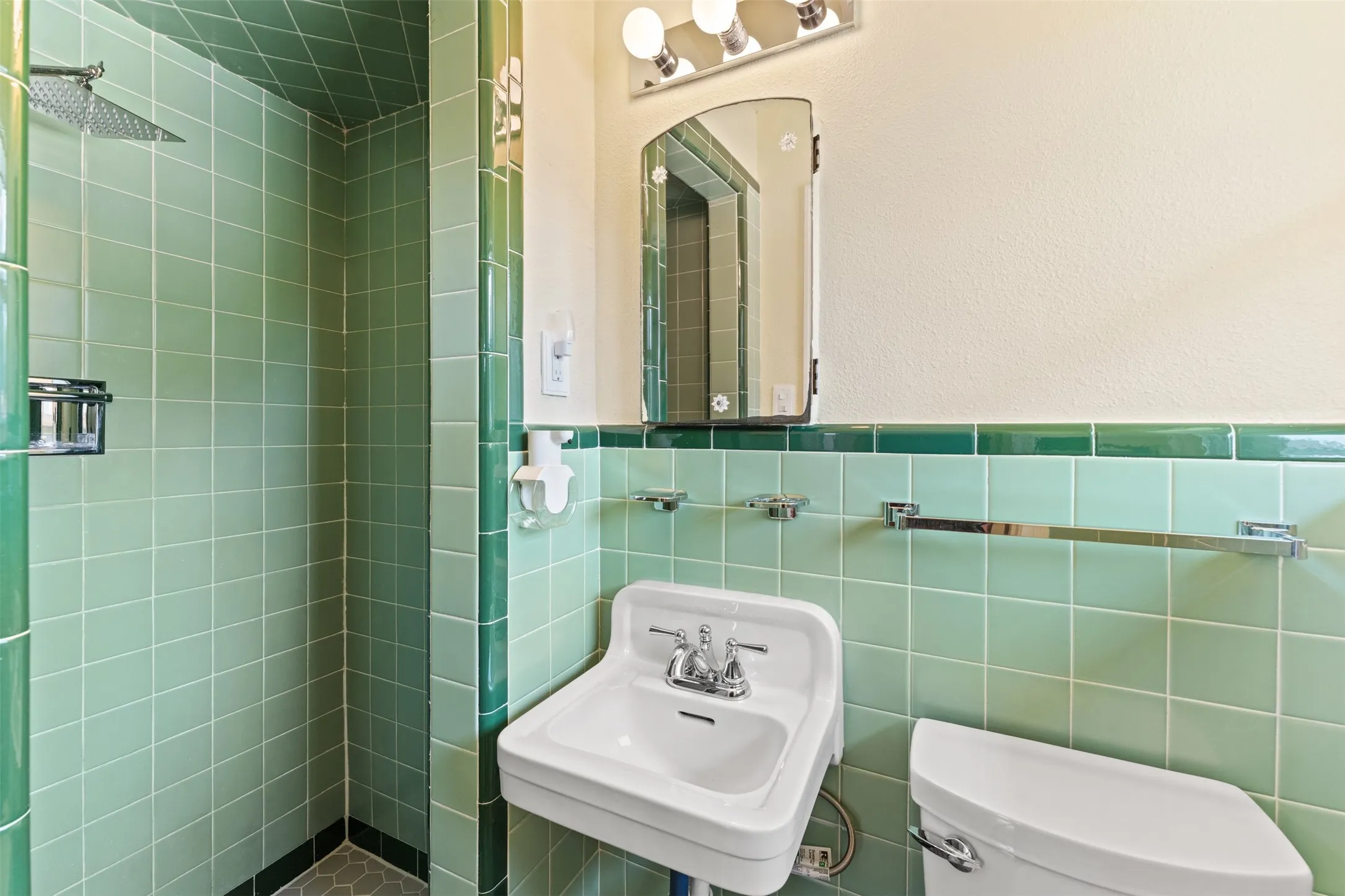 Bathroom featuring light green square tile walls with dark green accent trim, a built-in shower with a rainfall showerhead, a wall-mounted sink, and a toilet